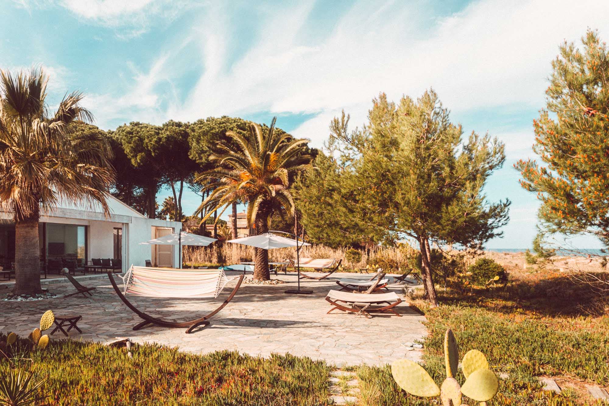 A sunny patio with hammocks and deckchairs is surrounded by palm trees and greenery. A modern house with a white exterior stands in the background under a blue sky dotted with fine clouds.