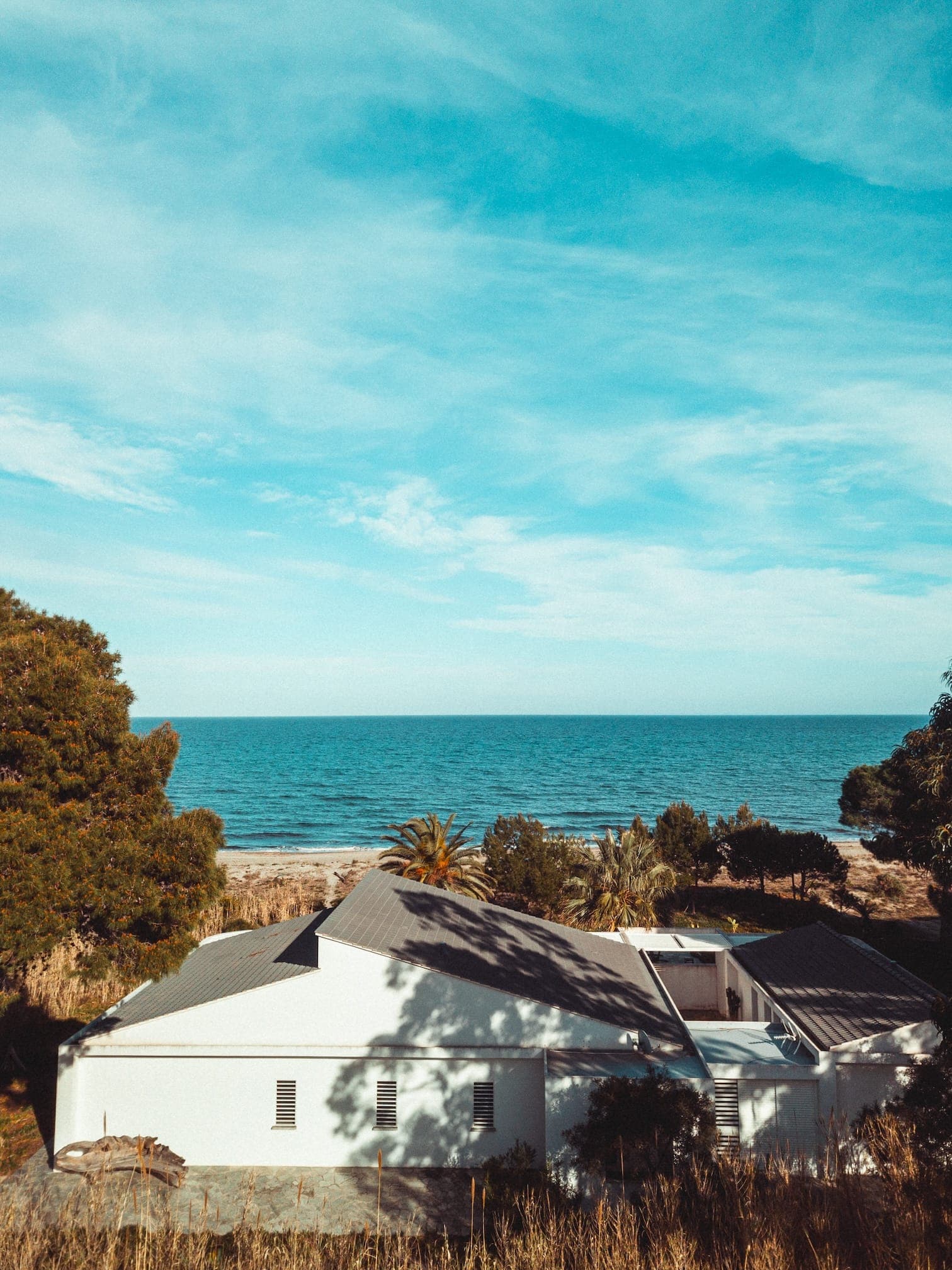 A white house with a gray roof is surrounded by trees, overlooking the ocean under a blue sky dotted with clouds. The grassy foreground leads to an unobstructed view of the sea and horizon.