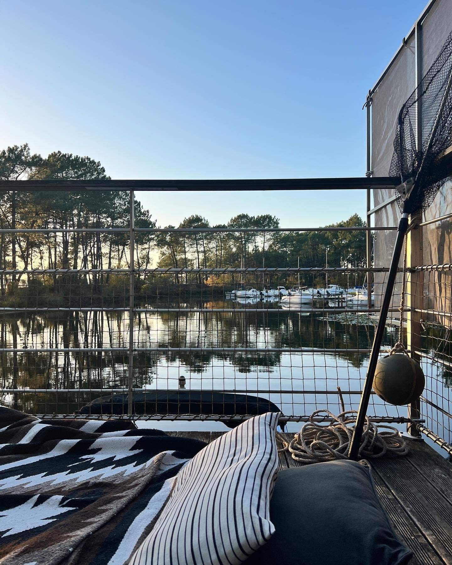 Vue depuis un bateau montrant un front de mer calme avec des arbres et des bateaux amarrés en arrière-plan. Une zone de pont comprend une couverture à motifs noirs et blancs, un oreiller rayé, une corde et une bouée, sous un ciel bleu clair.