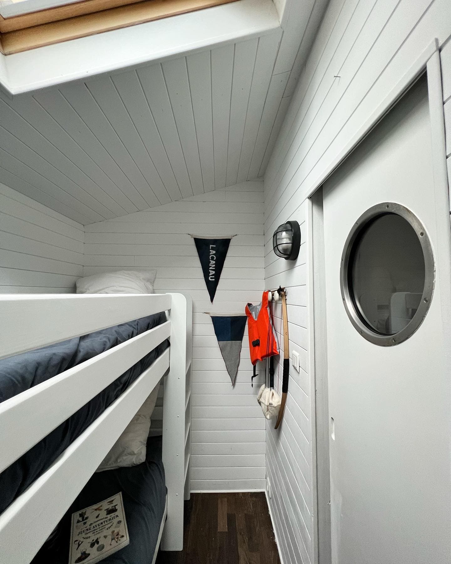 Petite chambre lambrissée de blanc avec lits superposés à gauche, avec literie grise. Les objets à thème nautique comprennent un fanion, un gilet de sauvetage et une fenêtre circulaire sur la porte. Une carte nautique repose sur la couchette inférieure. Parquet en bois foncé.