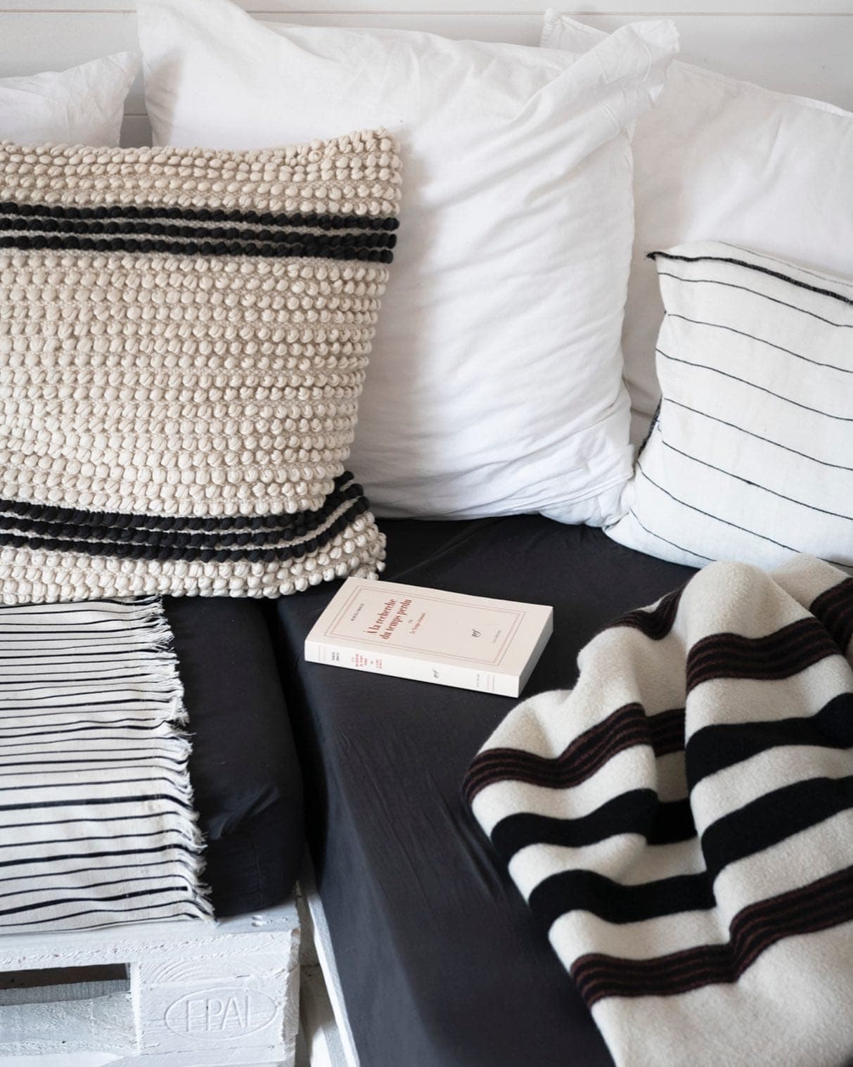 Un lit douillet avec des oreillers et une couverture à rayures blanches et noires. Un livre intitulé « Sept brèves leçons de physique » repose sur un drap foncé. Un motif rayé apparaît à la fois sur le jeté et sur l'oreiller tricoté. Des meubles en bois blanc sont en partie visibles.
