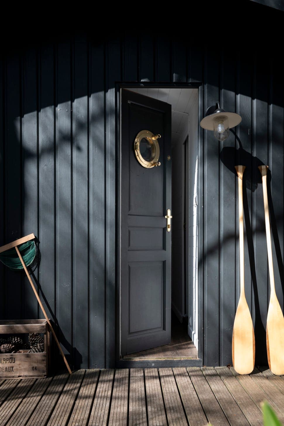 Une façade en bois sombre avec une porte ouverte dotée d'un hublot rond. Deux pagaies en bois sont appuyées contre le mur à côté d'un luminaire de style vintage. Une caisse avec un filet de pêche se trouve sur le pont, projetant des ombres depuis les arbres environnants.