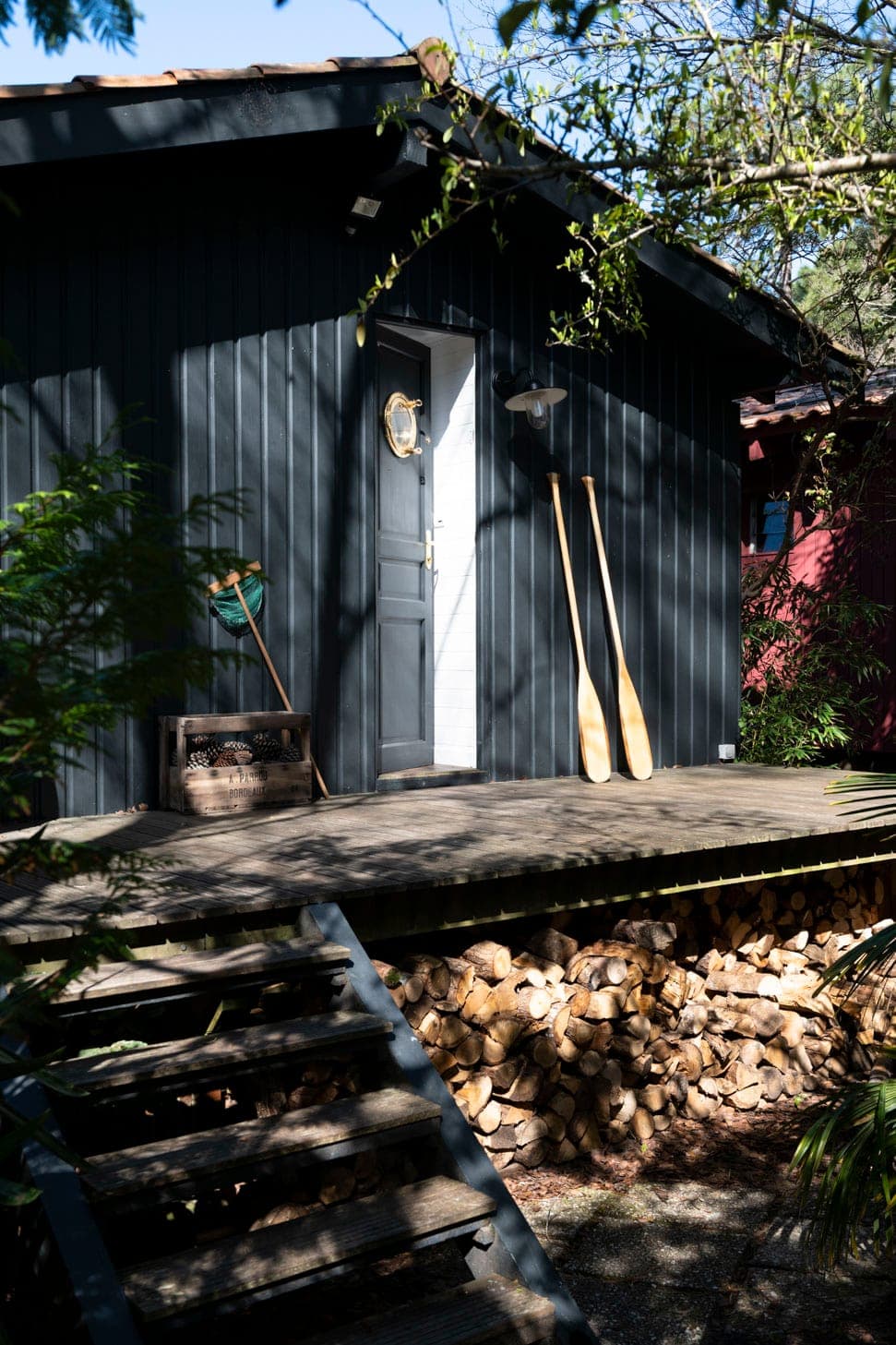 Une petite cabane en bois vert foncé avec une porte blanche est entourée d'arbres. Deux rames sont appuyées contre le mur de la cabane. Une pile de bois de chauffage coupé se trouve sous le porche surélevé, auquel on accède par des marches en bois. La lumière du soleil filtre à travers le feuillage.