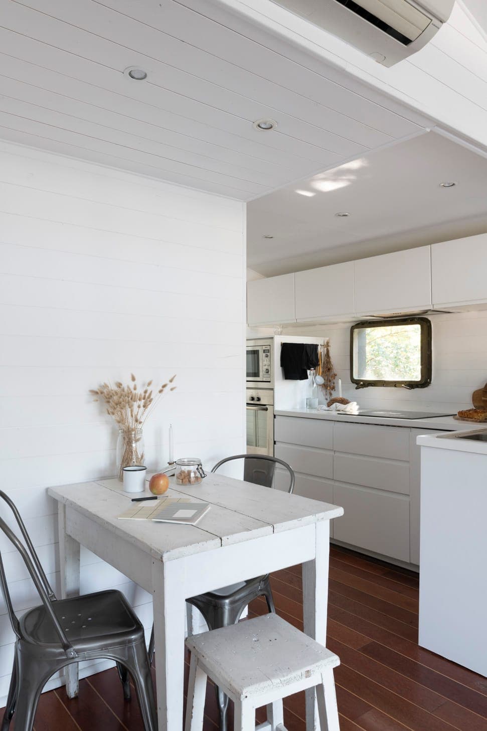 Une cuisine blanche minimaliste avec un sol en bois. Une petite table avec deux chaises en métal et un tabouret se trouve au premier plan. La table est ornée d'un arrangement de plantes séchées, d'une pomme et d'un livre ouvert. Une petite fenêtre offre une lumière naturelle.