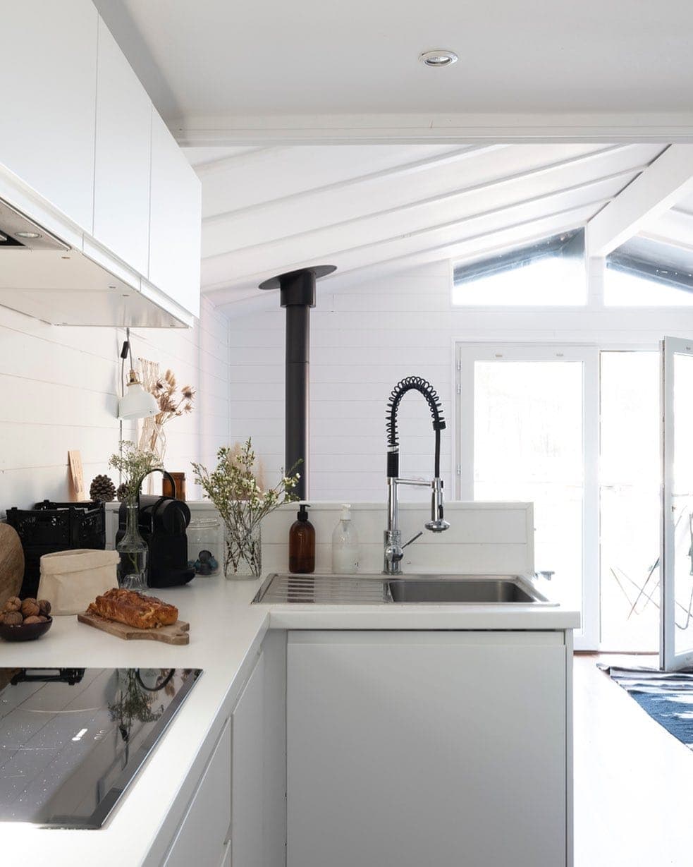Cuisine blanche moderne au design épuré, avec îlot central, évier en acier inoxydable et robinet à grand arc. Des touches de bois et des plantes ajoutent de la chaleur. Un poêle à bois et de grandes portes vitrées apportent de la lumière naturelle et une sensation d'espace.