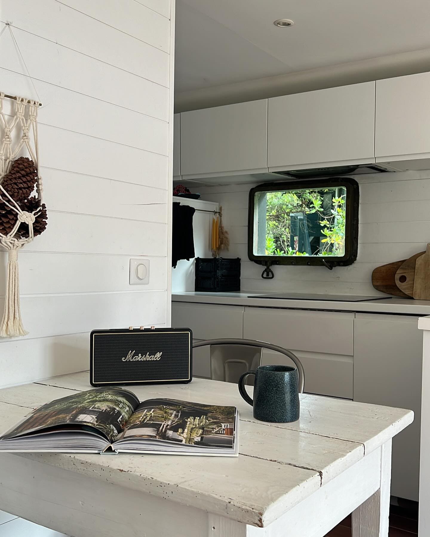 Une cuisine rustique et confortable avec des armoires blanches et une table en bois. Sur la table se trouvent un haut-parleur noir, un livre ouvert et une tasse sombre. Une fenêtre avec vue sur la verdure se trouve au-dessus du comptoir et une décoration suspendue est accrochée au mur.