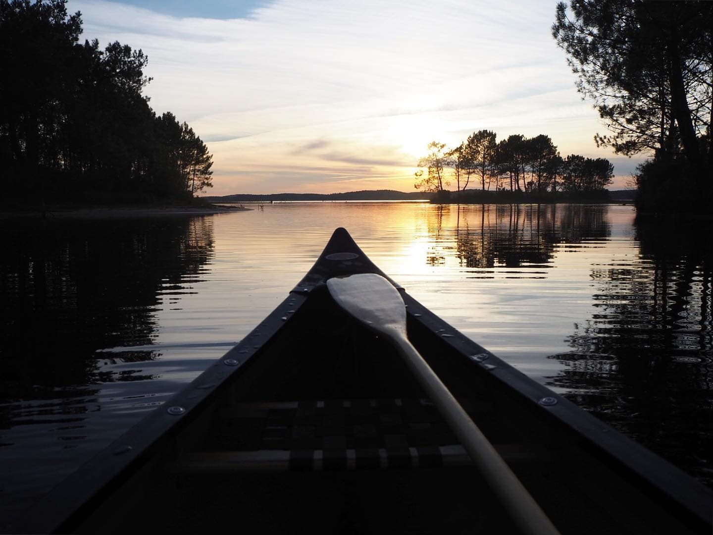 Un canot glisse sur une eau calme au coucher du soleil, entouré de silhouettes d'arbres et reflétant le ciel doré. Une pagaie repose sur le bord du canot tandis que l'horizon brille d'orange et de rose, créant une atmosphère sereine.