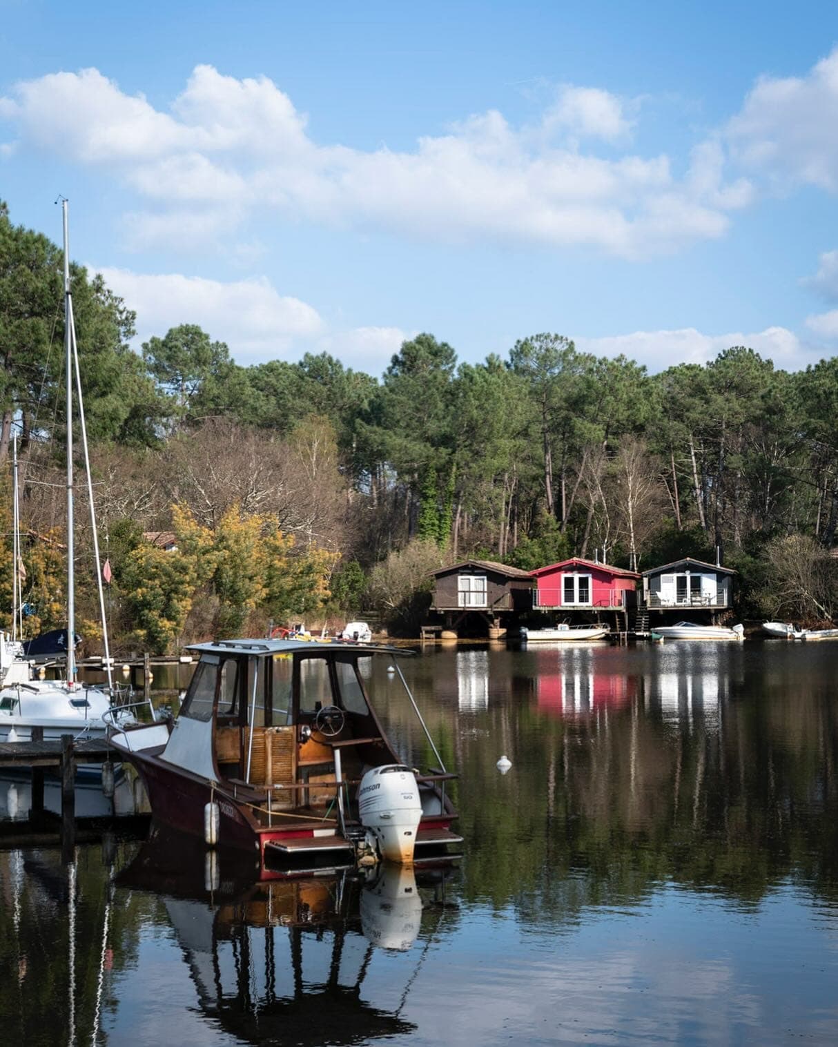 Une scène de lac paisible avec de petits bateaux amarrés au premier plan. De l'autre côté de l'eau, il y a de charmantes maisons en bois, l'une peinte en rouge et l'autre en blanc, entourées d'arbres luxuriants sous un ciel bleu clair.