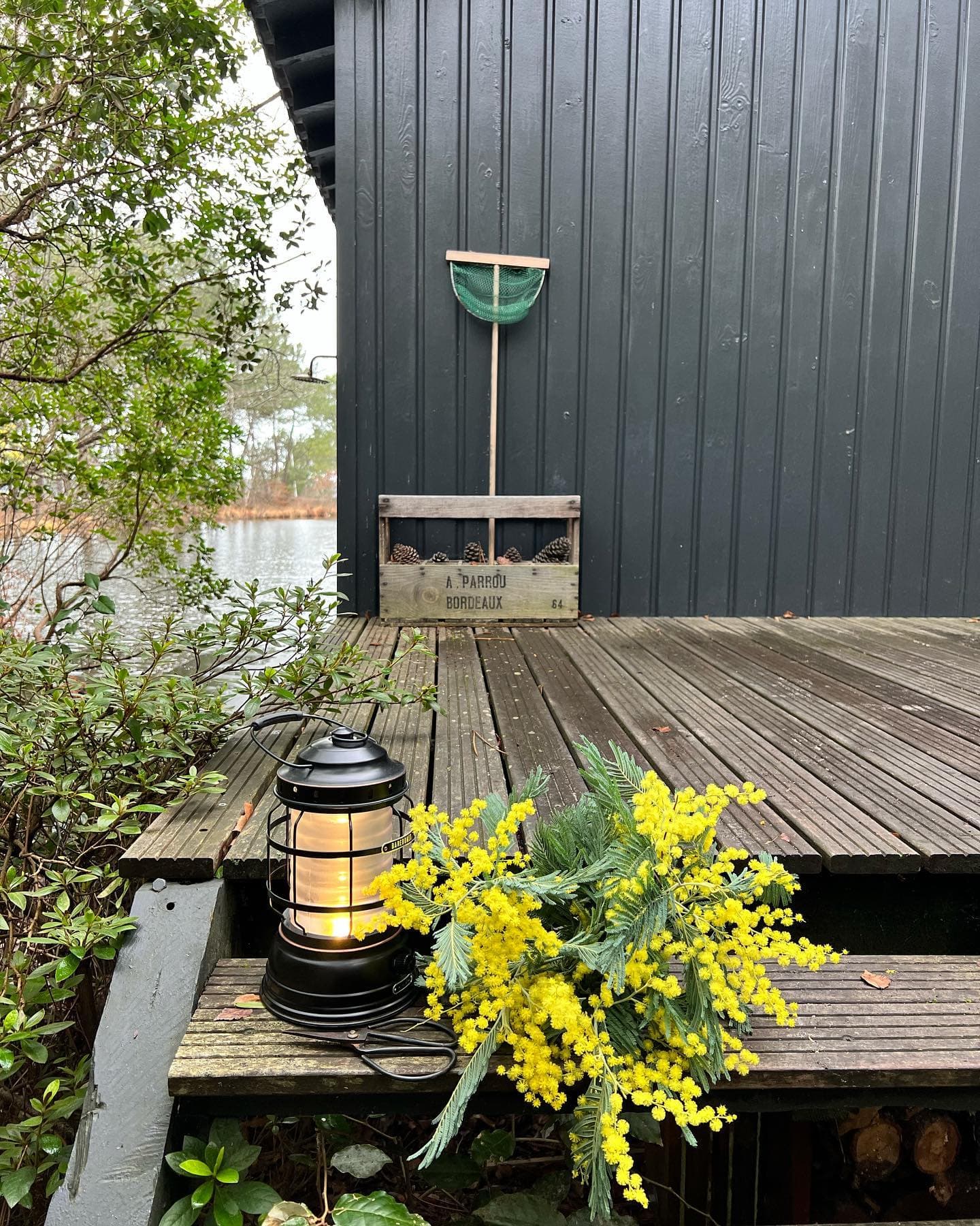 Une terrasse en bois avec une lanterne allumée et un bouquet de fleurs jaunes. Une caisse rustique avec un outil de jardinage est contre un mur gris foncé. La verdure entoure la zone et un plan d'eau est visible en arrière-plan.