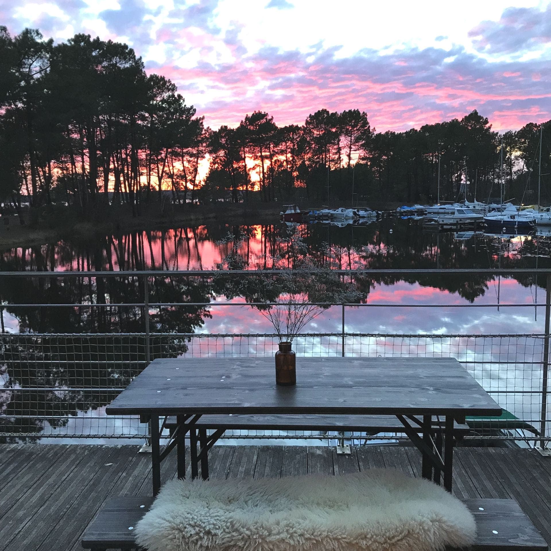 Une table de pique-nique en bois avec un plaid en fourrure est posée sur une terrasse surplombant un lac calme au coucher du soleil. Le ciel est rempli de teintes roses et violettes, et les arbres se découpent en silhouette sur les couleurs vives. Des bateaux sont amarrés de l'autre côté du lac.