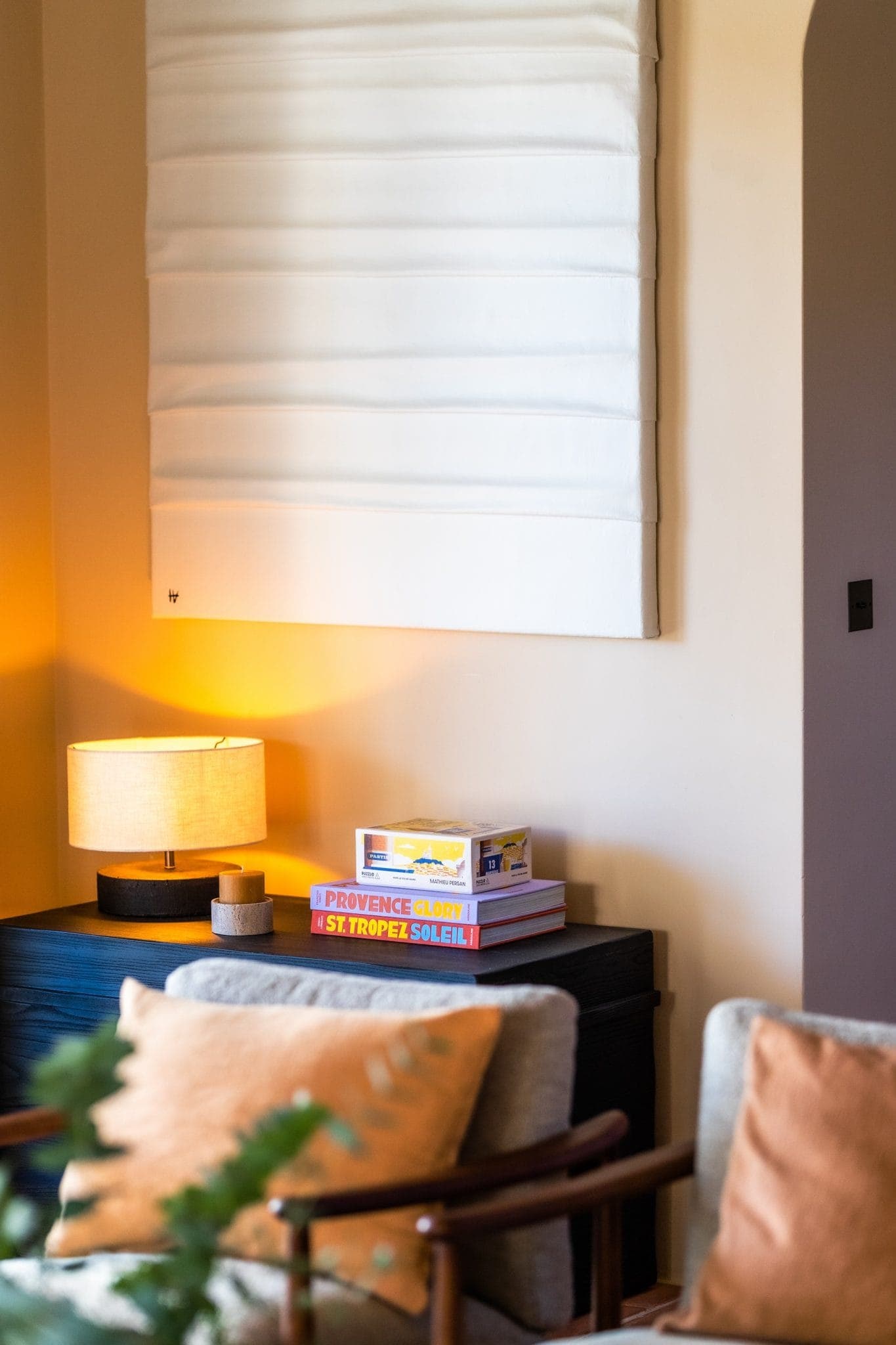 A cozy room corner features a black table with a beige lamp and stacked colorful books. Softly lit by warm lighting, the space includes a white wall hanging and cushioned chairs, enhancing the cozy atmosphere.