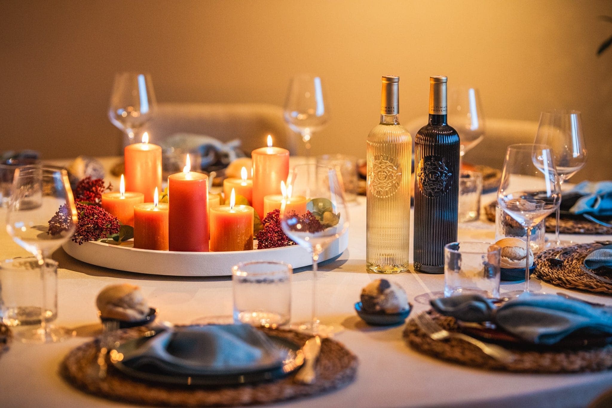 A warmly lit dinner table is set with candles, two bottles of wine, and glassware. Each place setting includes plates with blue napkins and decorative items. The candles create a cozy atmosphere.
