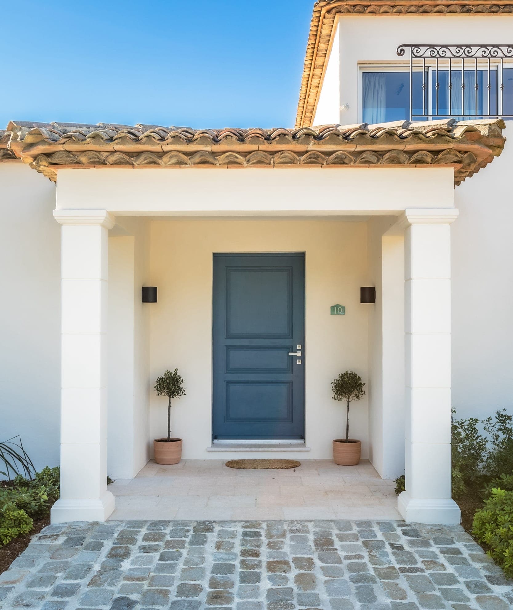 Front entrance of a house featuring a blue door set between two white columns. Two small potted plants flank the doorway. The roof has terracotta tiles, and cobblestones pave the walkway. A small balcony with a metal railing is visible above.