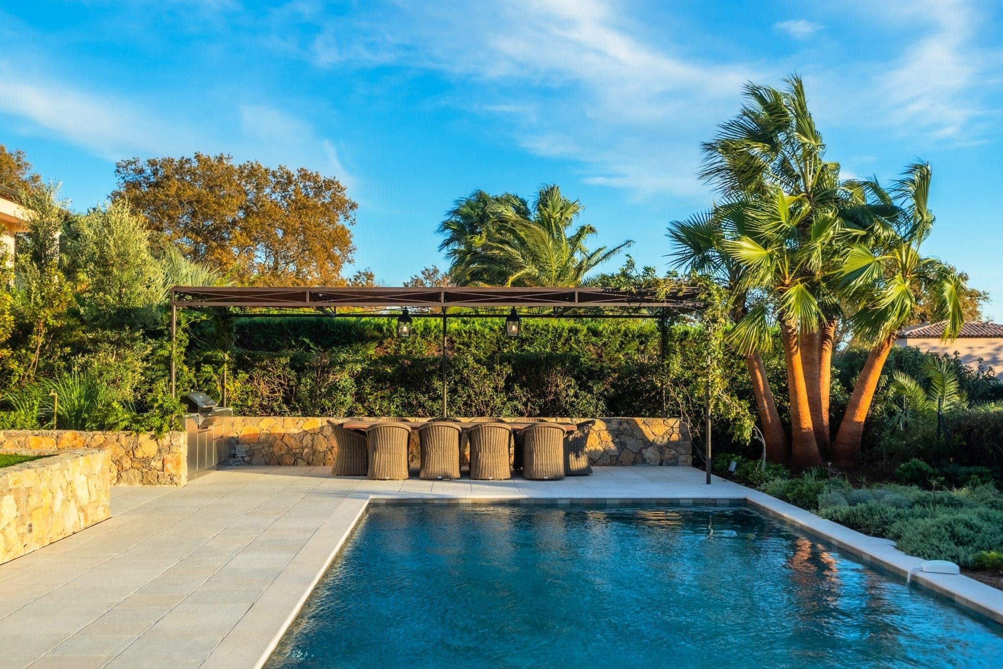 A serene outdoor pool area with clear blue water, surrounded by lush greenery and palm trees. There's a covered seating area with wicker chairs and a stone wall in the background under a bright blue sky.