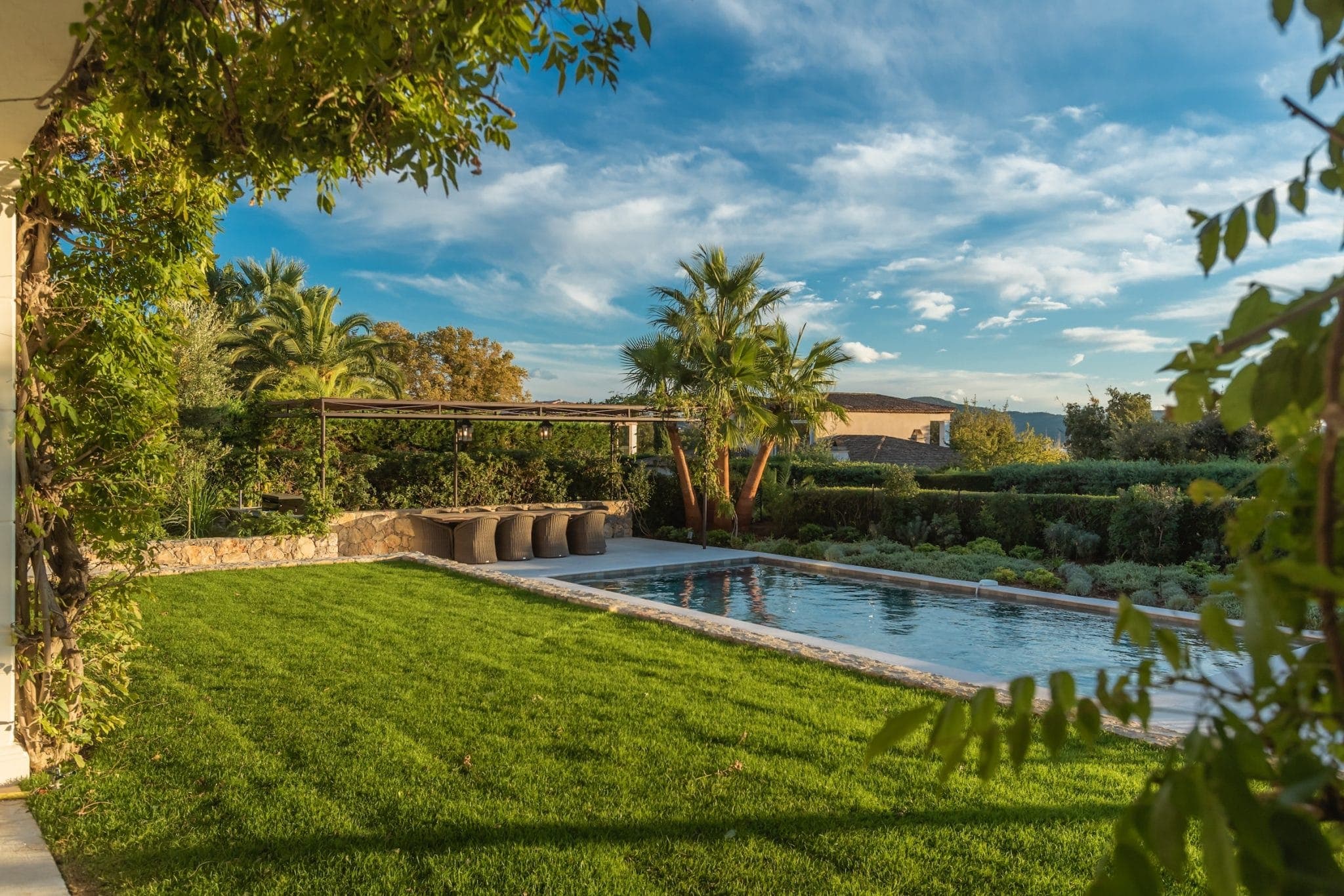 A vibrant garden with a neatly trimmed lawn surrounds a rectangular swimming pool. Palm trees and shrubs line the area, with a covered patio and dining set on one side. The sky is blue with scattered clouds, adding a serene atmosphere.