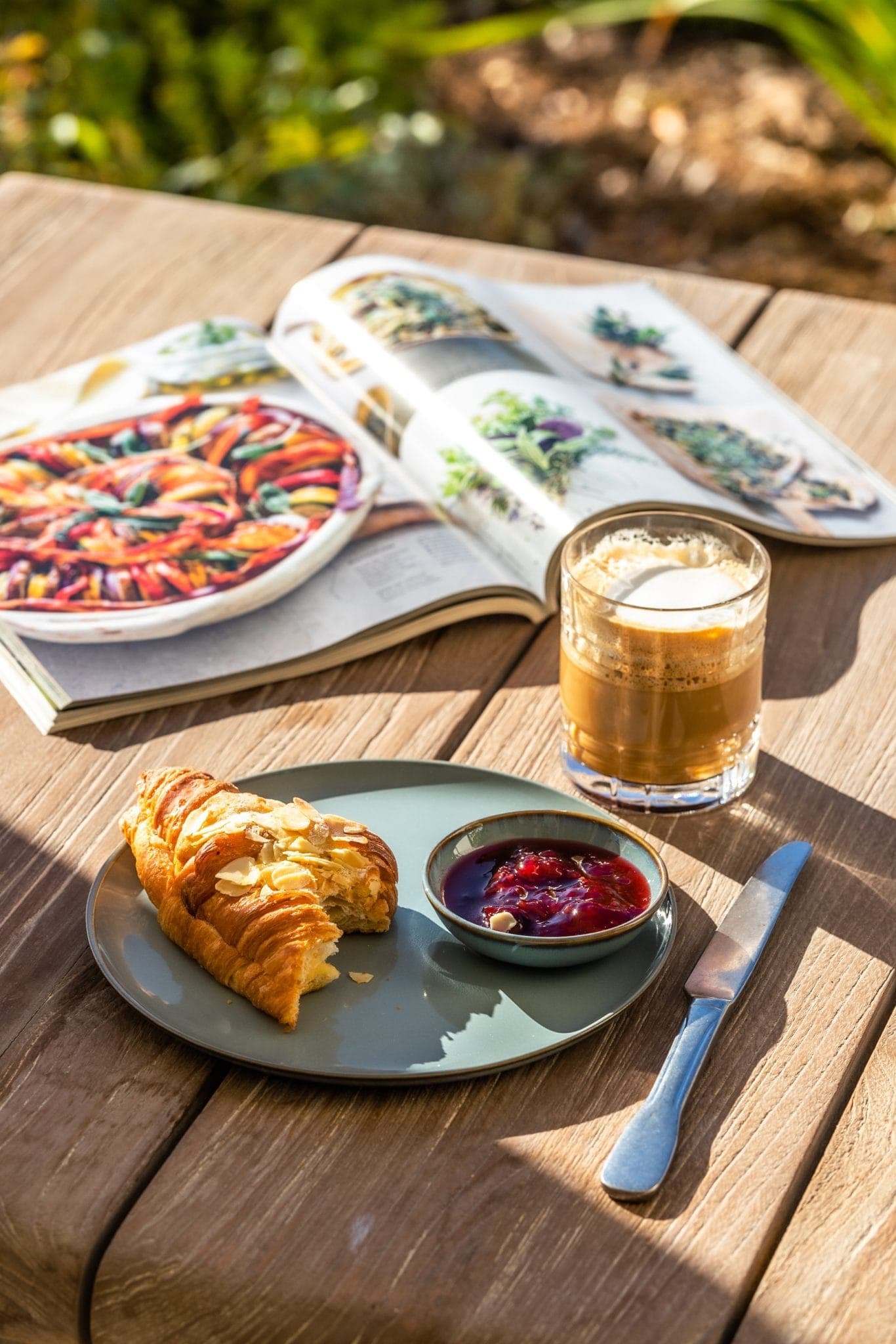 A croissant and a small dish of red jam sit on a green plate on a wooden table. A knife rests beside it. A glass of coffee and an open, colorful cookbook are also on the table, with greenery in the background.