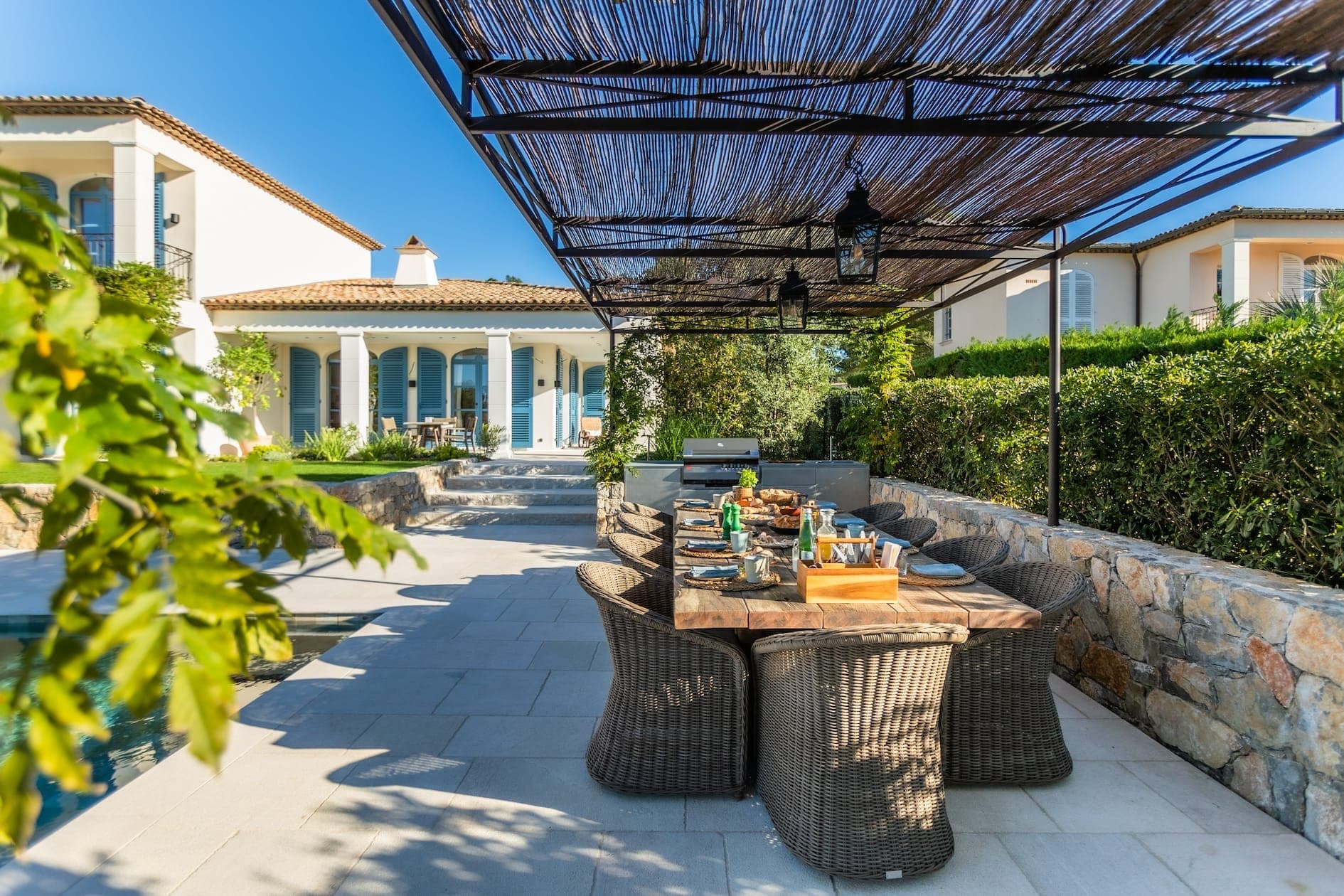 Outdoor dining area with a wooden table set for a meal, surrounded by wicker chairs. A pergola with a woven canopy provides shade. In the background, a Mediterranean-style house with white walls and terracotta roof tiles is visible. Lush greenery surrounds.