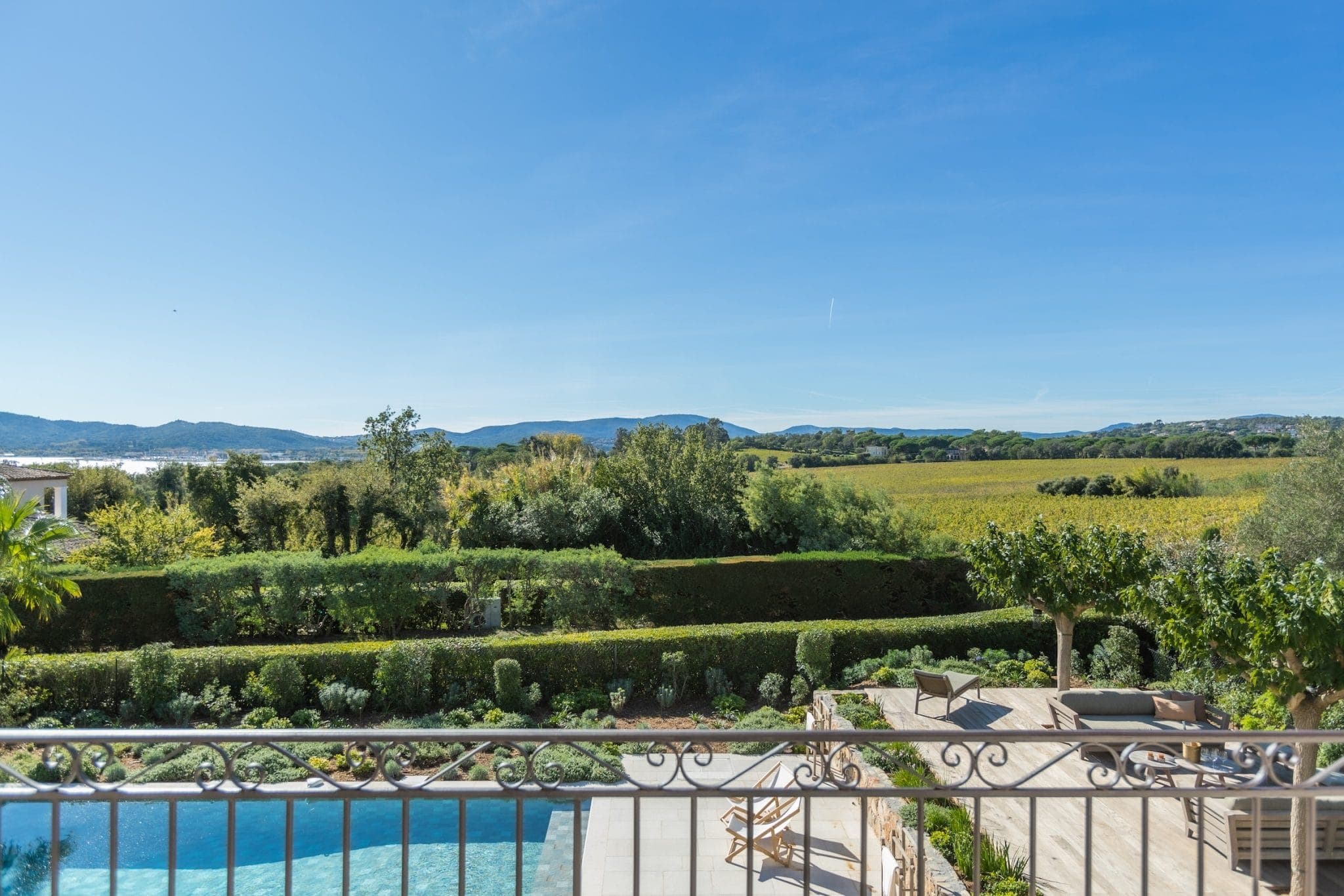 View from a balcony overlooking a serene landscape with lush greenery, trees, and distant hills under a clear blue sky. Below, a patio with outdoor furniture is adjacent to a swimming pool, bordered by well-maintained gardens and hedges.