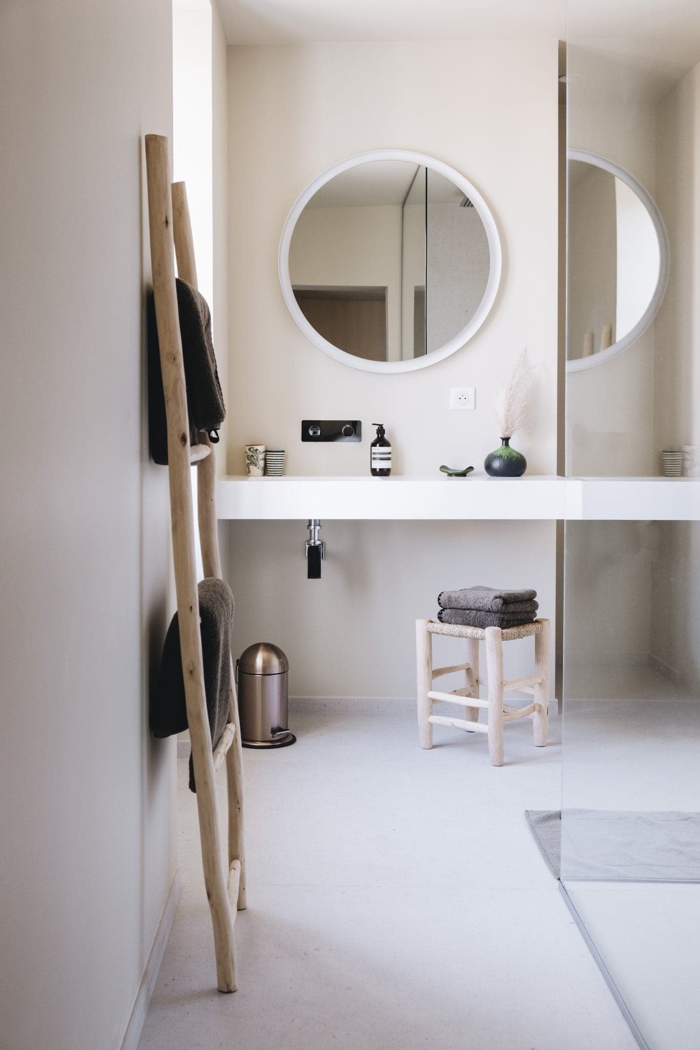 Minimalist bathroom with a circular mirror above a white floating vanity. A wooden ladder holds towels, and a small wooden stool with stacked towels sits nearby. A potted plant adds a touch of greenery. A glass shower screen is visible on the right.