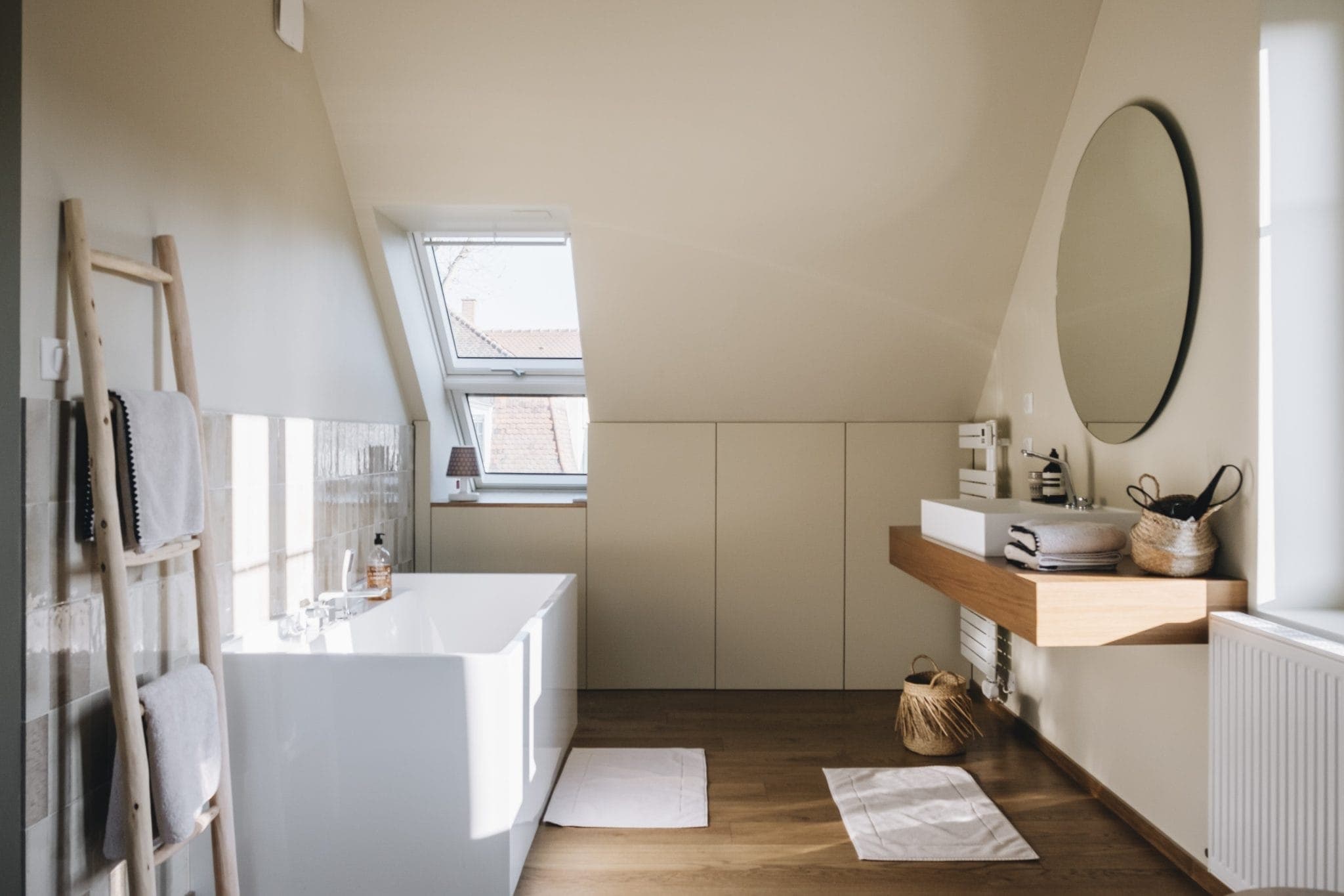 A modern bathroom with a slanted ceiling features a white bathtub, a wooden vanity with a circular mirror, and an open window letting in natural light. Towels hang on a ladder rack, and a wicker basket sits on the wooden floor.