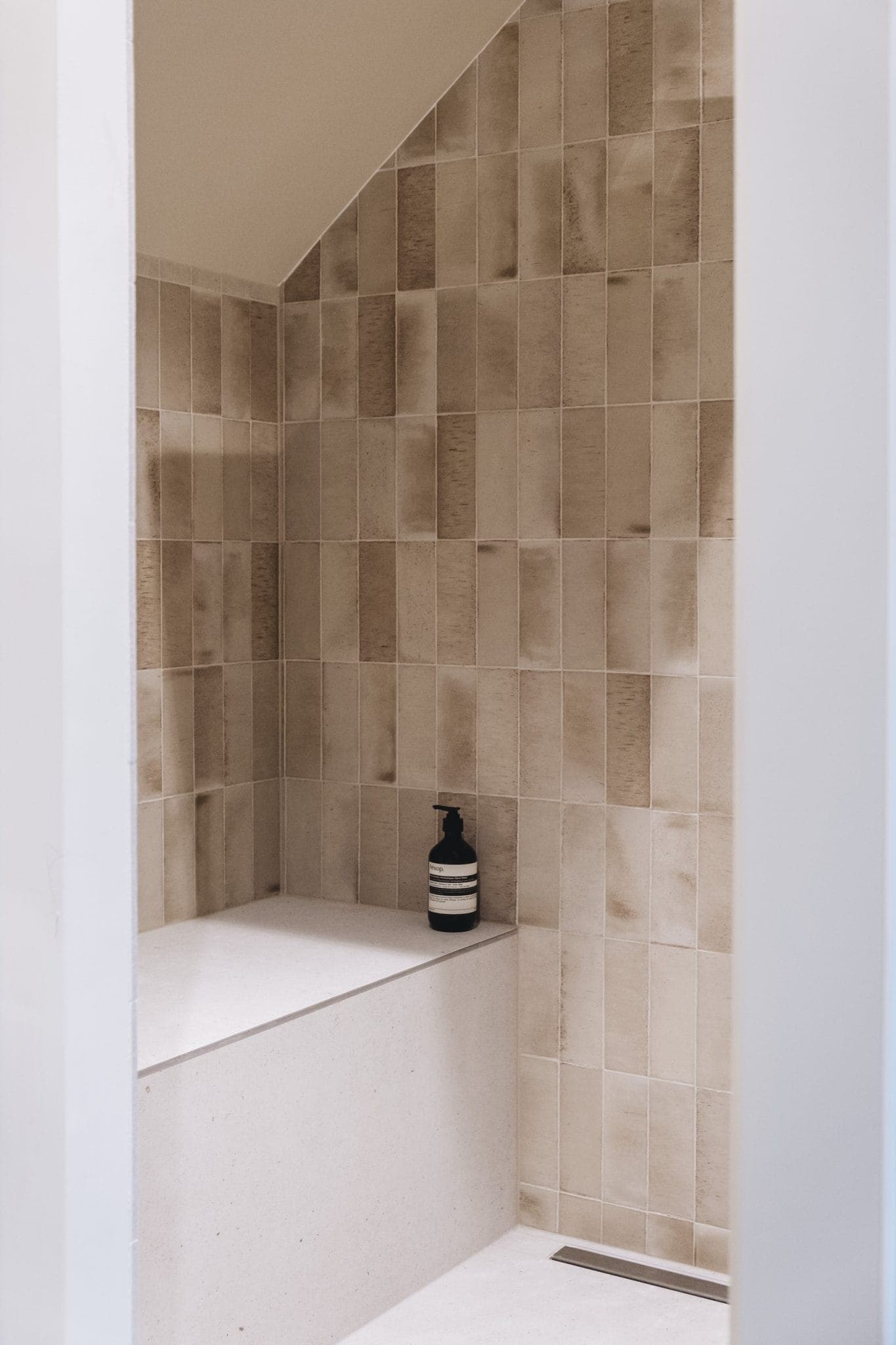 A minimalist shower with beige tiled walls and a built-in white bench. A black and white striped soap dispenser sits on the bench, adding a modern touch to the neutral-colored space.