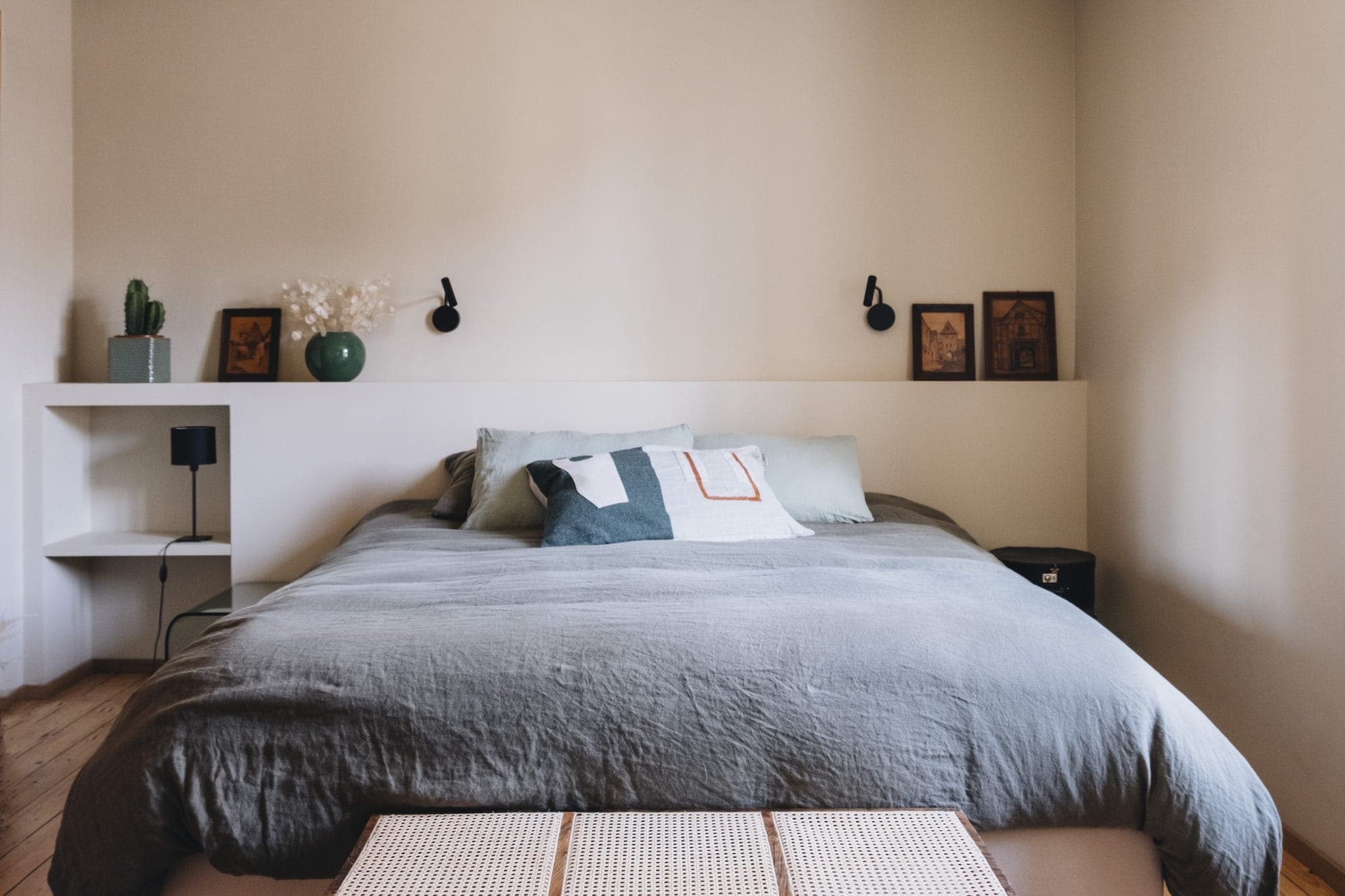 A cozy bedroom with a neatly made bed covered in gray linens and decorative pillows. The wall features two small framed pictures and two wall-mounted reading lights. A shelf on the left holds a vase with flowers and a small cactus.