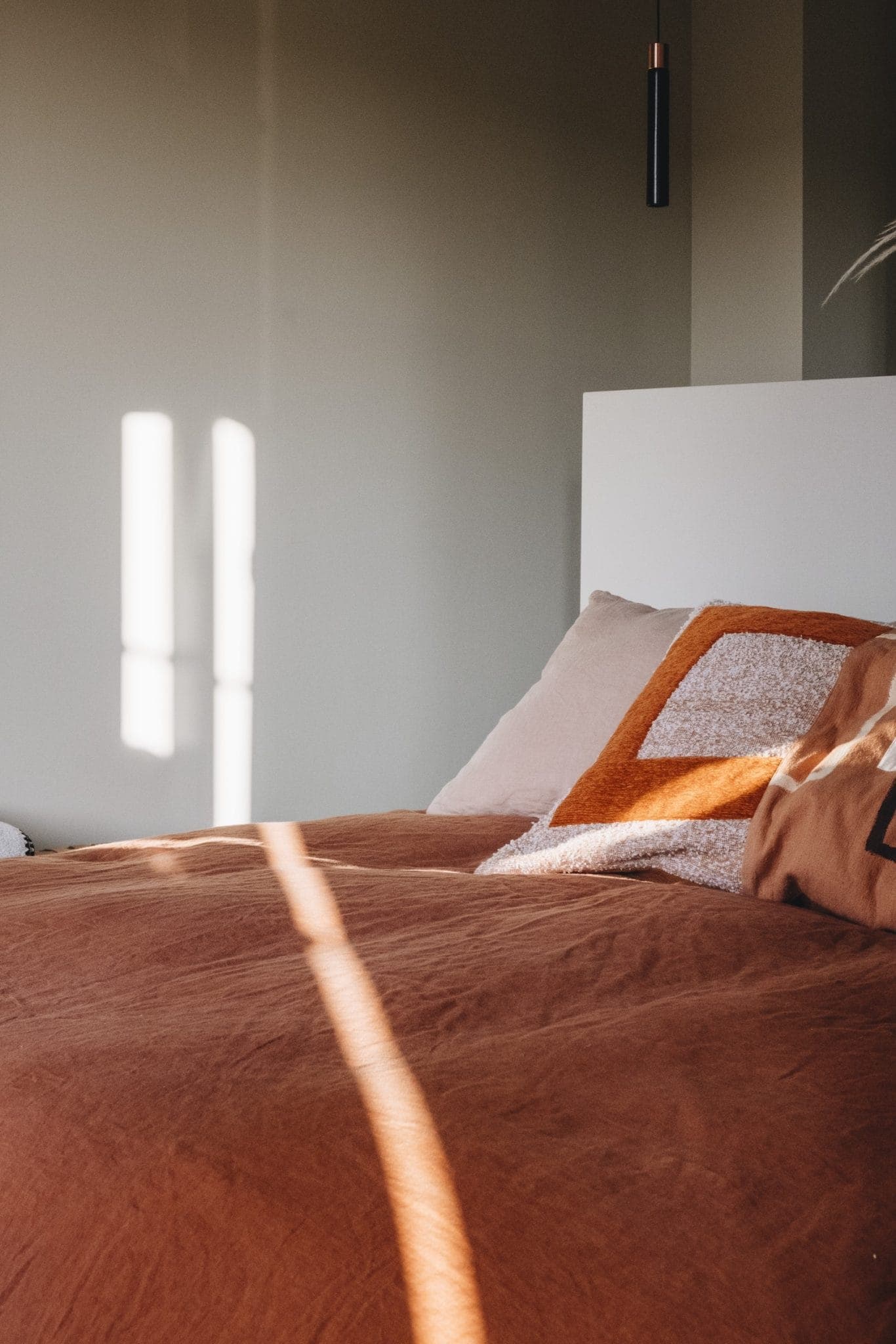 A cozy bedroom scene with sunlight streaming through a window, casting shadows on a bed. The bed is made with rust-colored linens and pillows, including one with an orange and white geometric pattern. The walls are a soft green color.