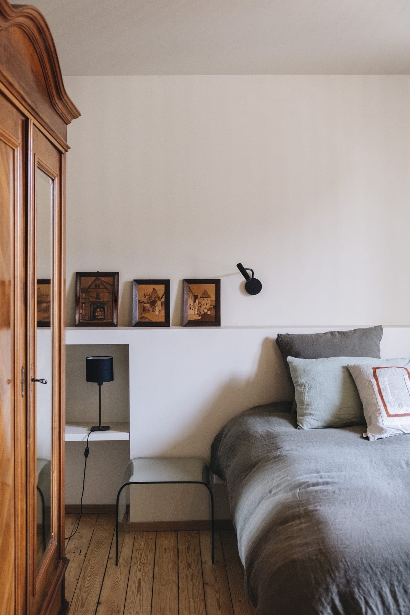 A cozy bedroom featuring a wooden wardrobe on the left, a neatly made bed with gray and white pillows on the right, a minimalist wall lamp, and small framed pictures above a white shelf with a black lamp and a glass side table.