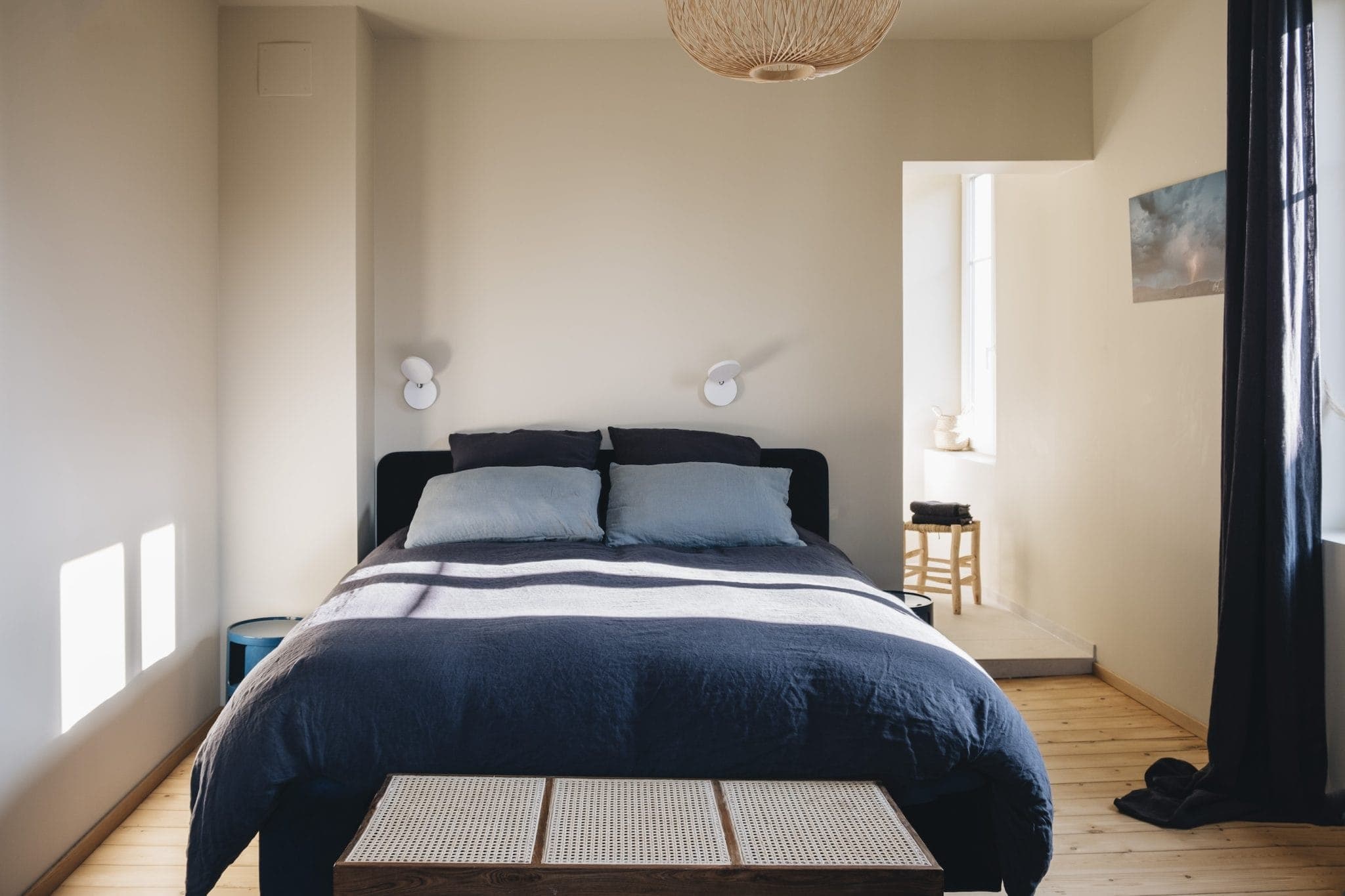 A minimalist bedroom with a double bed featuring dark bedding and light pillows. Two wall-mounted lamps are above the bed. The room has wooden flooring, a window with dark curtains, a round light fixture on the ceiling, and a small bench at the base of the bed.