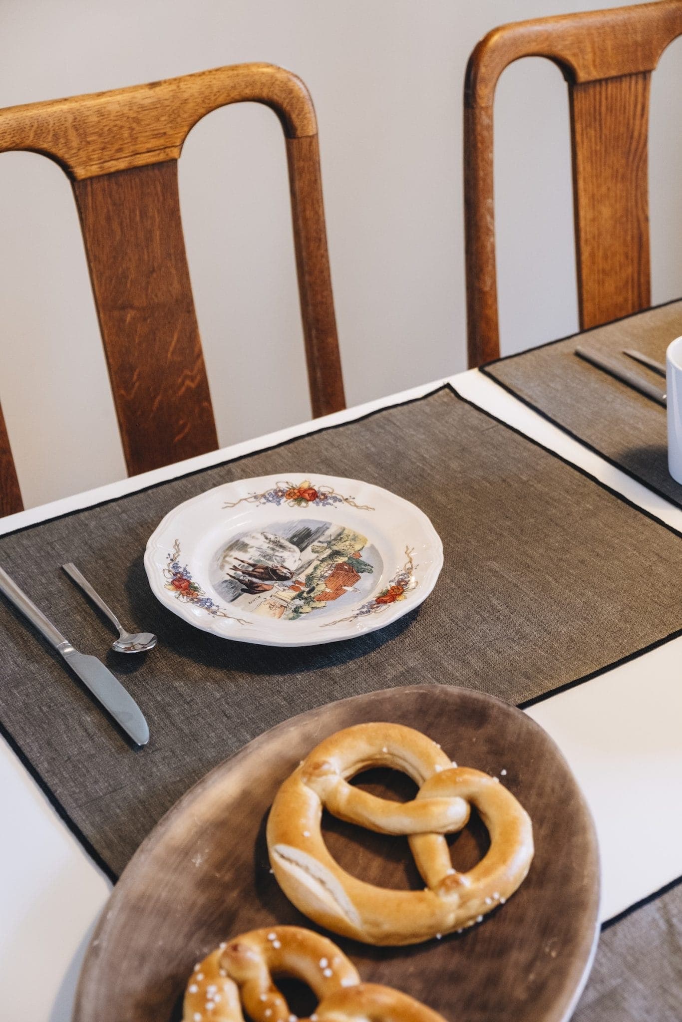 A dining table setting with two wooden chairs. A decorative plate with a scenic design is placed on a gray placemat with a fork and spoon. In the foreground, a wooden tray holds two soft pretzels.