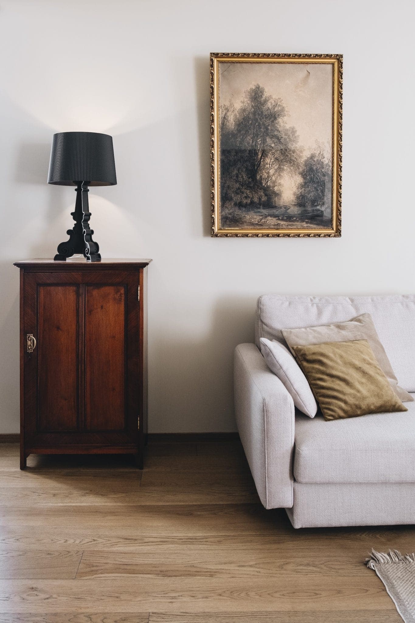 A cozy living room scene featuring a soft beige sofa with cushions, a dark wooden side cabinet, and a black table lamp. A framed landscape painting hangs on the light-colored wall, and the floor is wooden.