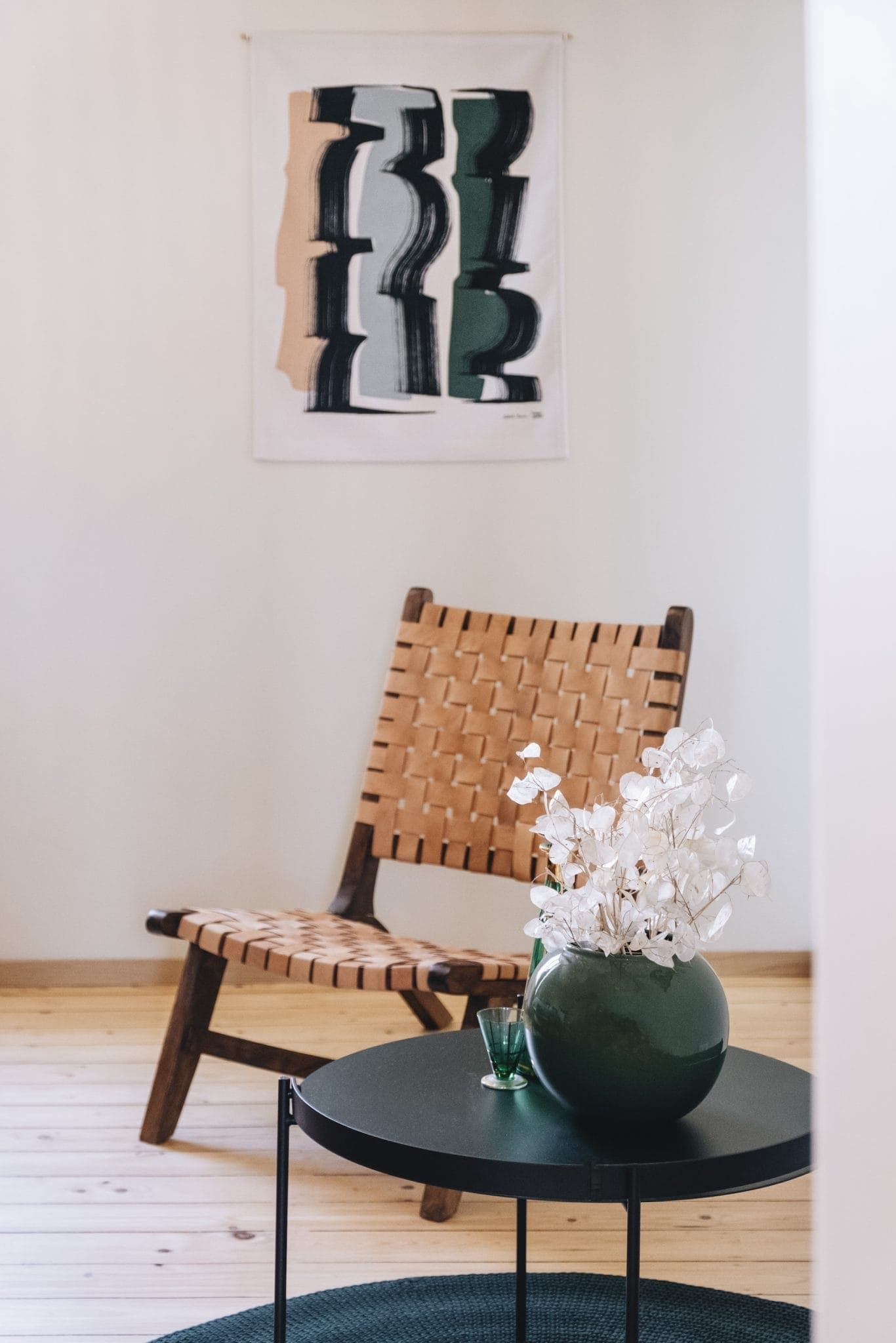 Minimalist room with a woven wooden chair against a light wall with abstract art. In the foreground, a round black table holds a green vase with white flowers, set on a wooden floor with a small round rug.