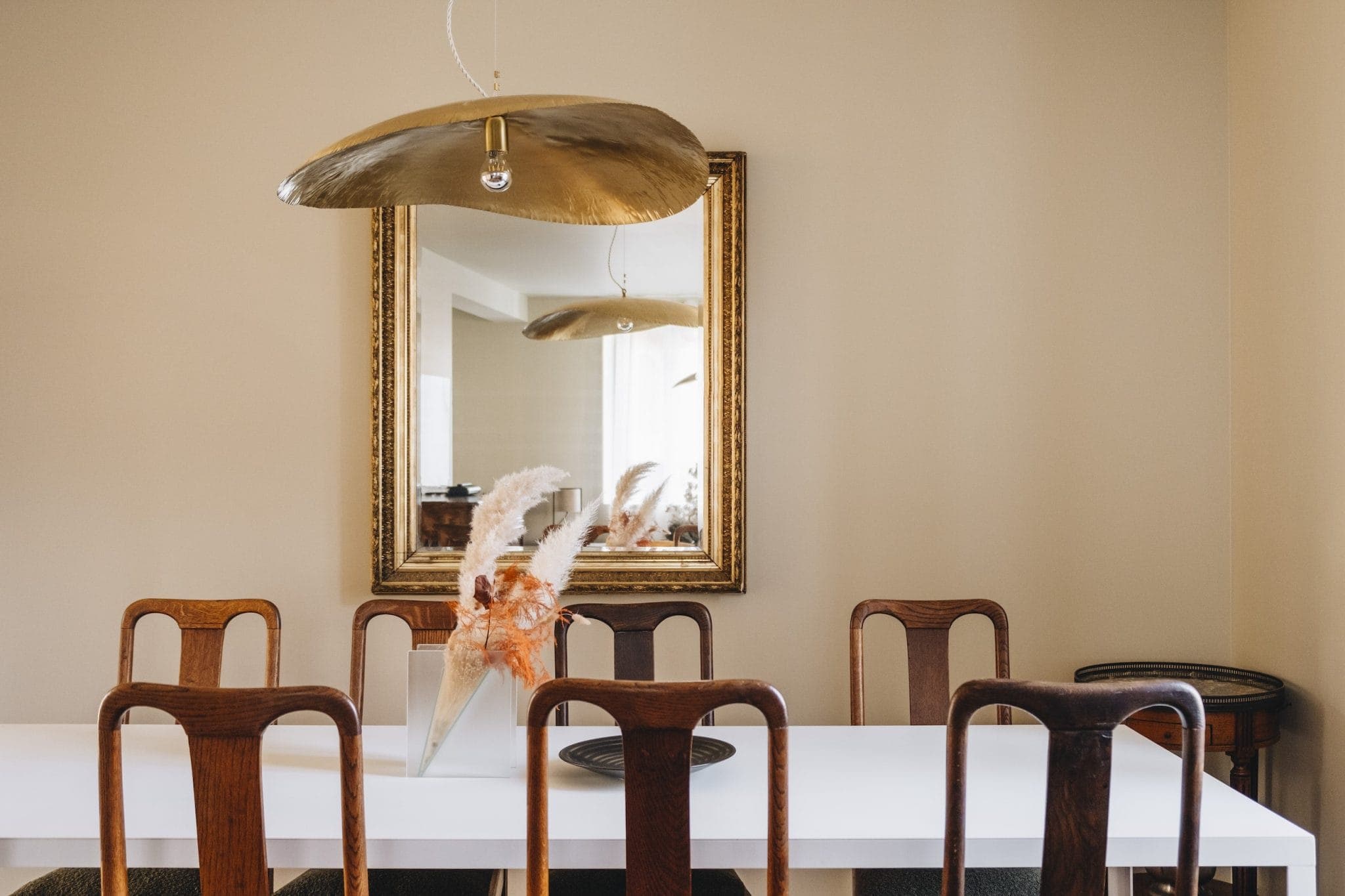 A dining room with a white table and six wooden chairs. A vase with pampas grass serves as a centerpiece. A large mirror in a gold frame hangs on the beige wall, reflecting a lamp with a large, gold, dome-shaped shade.
