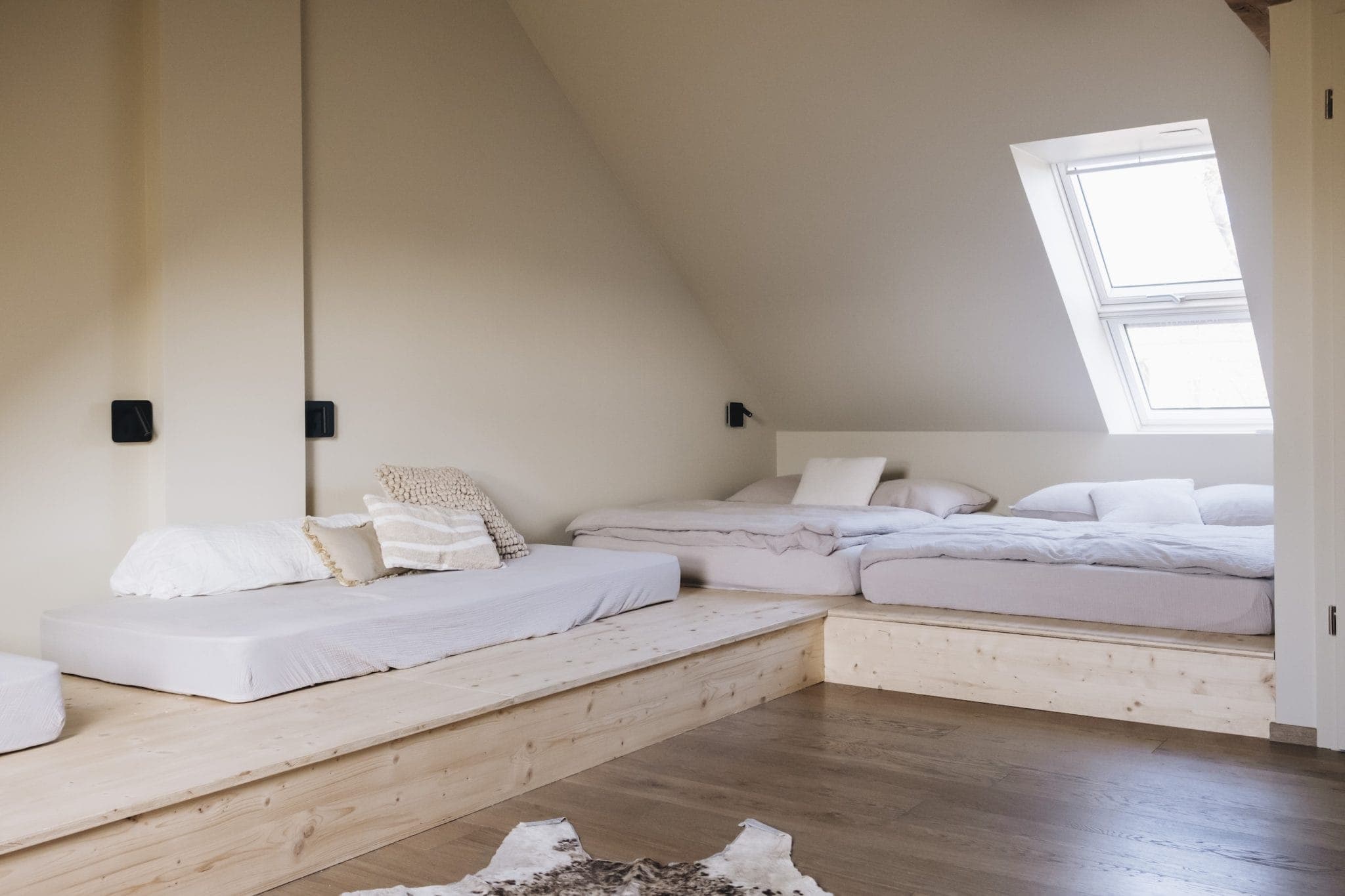 A cozy attic bedroom with two platform beds, each covered with light bedding. The sloped ceiling features a skylight, allowing natural light to fill the space. The room's wooden flooring and neutral color palette create a warm, inviting atmosphere.