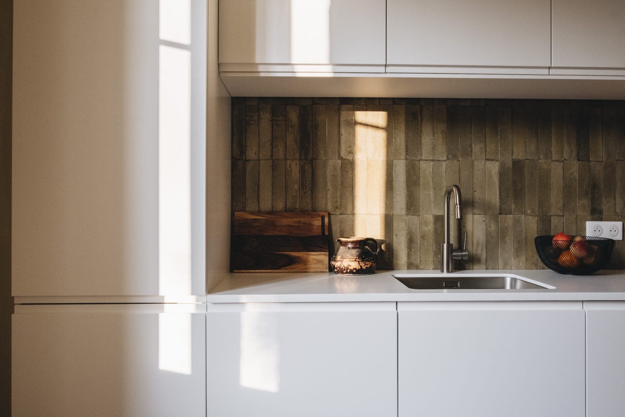 A modern kitchen with white cabinets, a sleek faucet, and a stainless steel sink. Sunlight casts shadows on the textured brown backsplash. A fruit bowl with oranges and a wooden cutting board add warmth to the minimalist design.