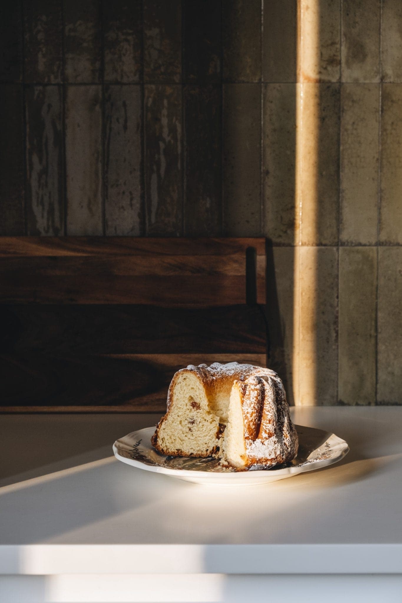 A partial bundt cake on a white plate sits on a light-colored surface. The cake is dusted with powdered sugar, and sunlight illuminates the scene, casting shadows on the tiled wall in the background.