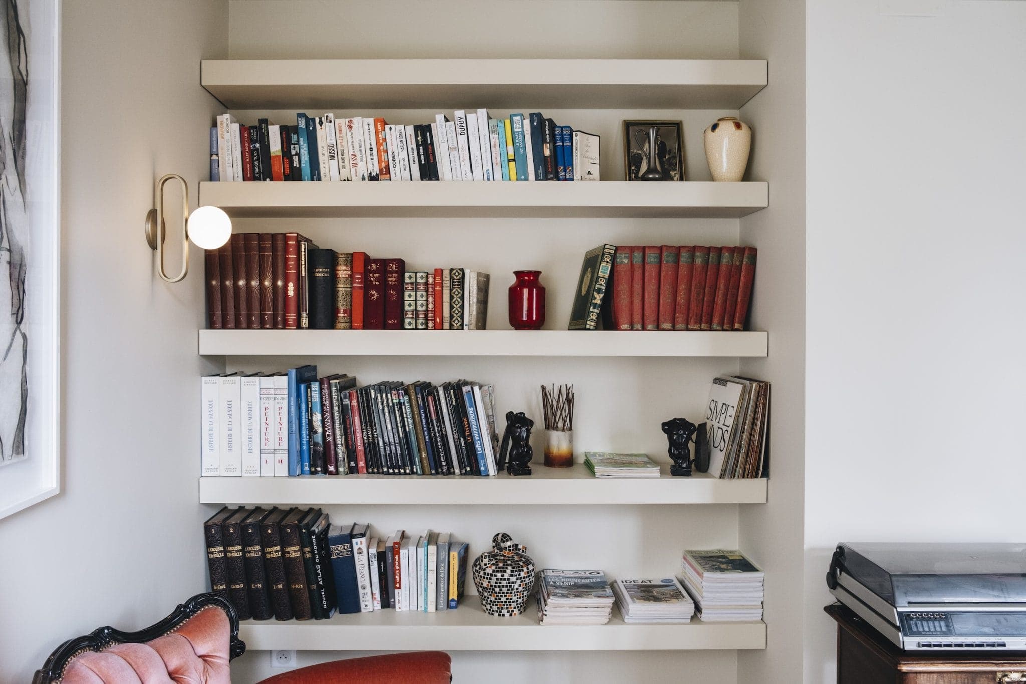 A wall-mounted bookshelf with various books, ornaments, and decorative items. The shelves hold a mix of hardcover and paperback books, with jars and small sculptures interspersed. A pink armchair and a printer are partially visible in the room.