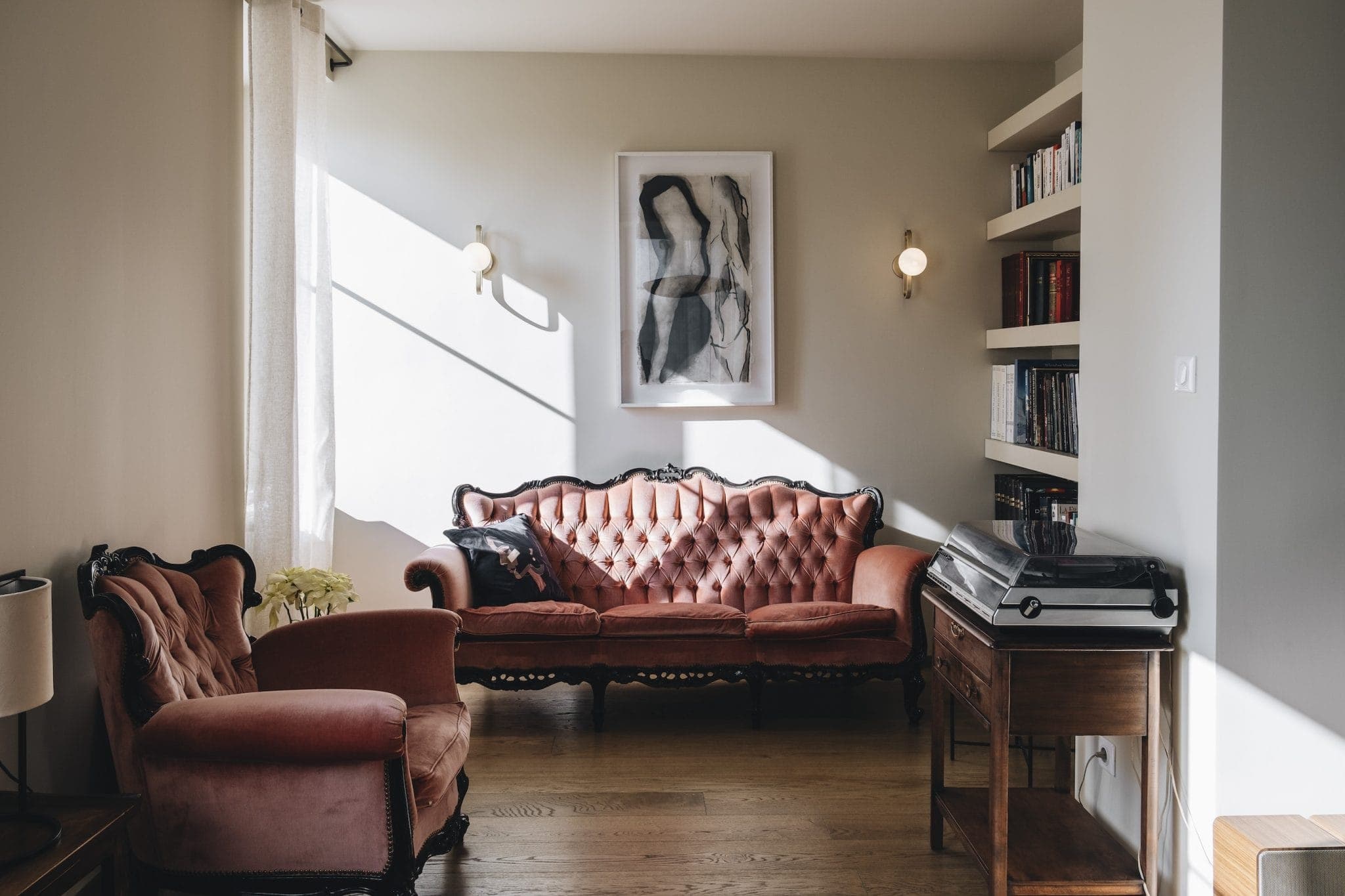 A cozy living room with a vintage pink tufted sofa and armchair, a wooden side table with a record player, and bookshelves. A framed abstract artwork hangs on the wall. Natural light streams in through a window with sheer curtains.