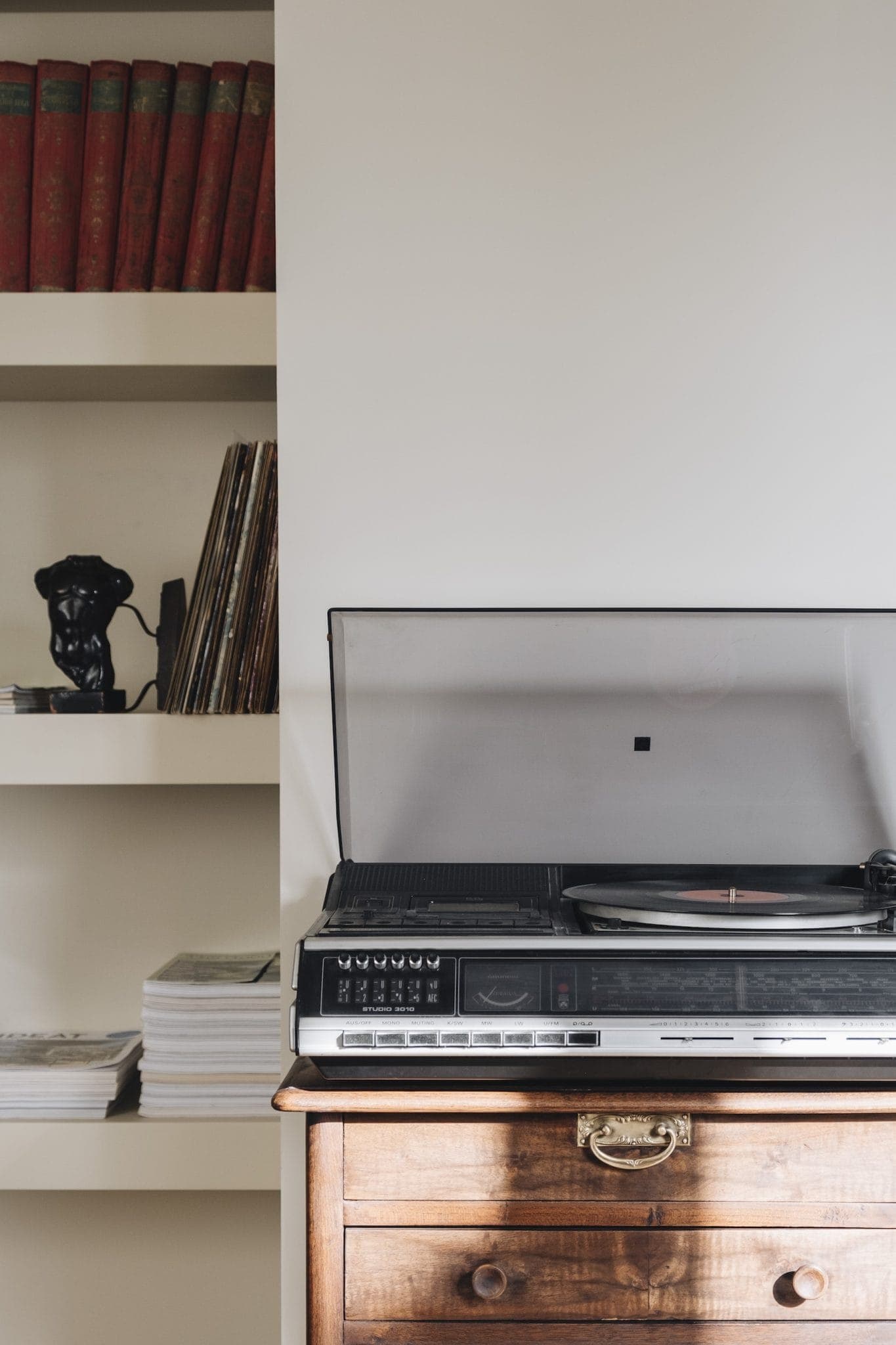 A vintage record player with a clear lid sits on a wooden dresser. To the left, a shelf holds books, vinyl records, and a decorative bust. The scene has a nostalgic, classic ambiance.