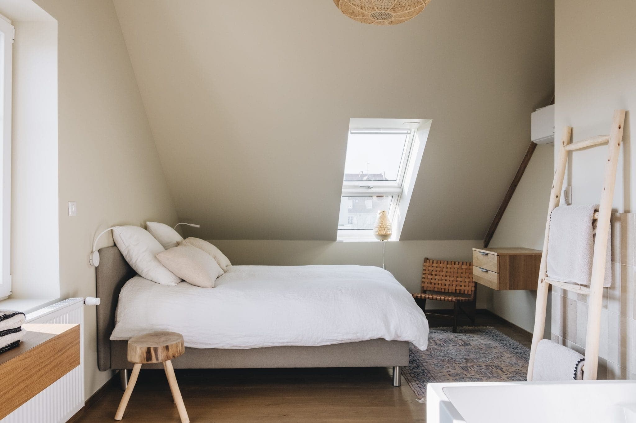 A cozy bedroom with sloped ceilings, featuring a neatly made bed with white linens, a small wooden stool, and a woven chair by a window. A rug covers part of the wooden floor, and a hanging light fixture adds to the warm ambiance.