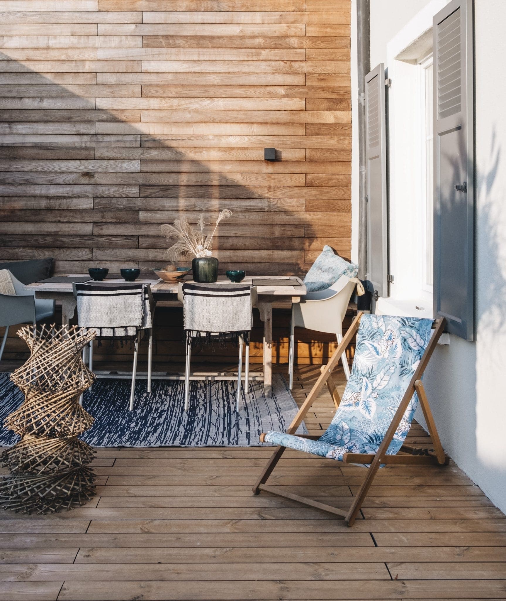 Outdoor patio with wooden decking and a slatted wooden wall. Features a dining table with white chairs, a decorative rug, and a floral deck chair. Potted plants and pillows add a cozy touch, and sunlight filters through an open window shutter.
