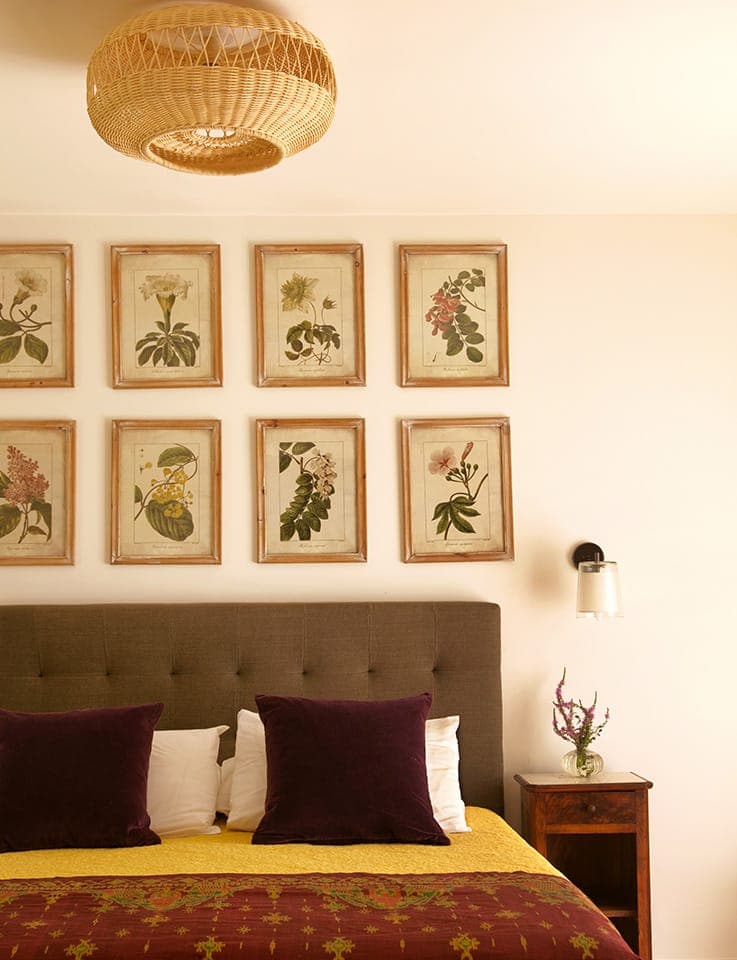 Une chambre douillette avec un lit aux coussins violet foncé et une housse à motifs jaune et bordeaux. Au-dessus du lit se trouvent neuf gravures botaniques encadrées. Un luminaire en osier et une petite table de nuit avec un vase de fleurs complètent le décor.