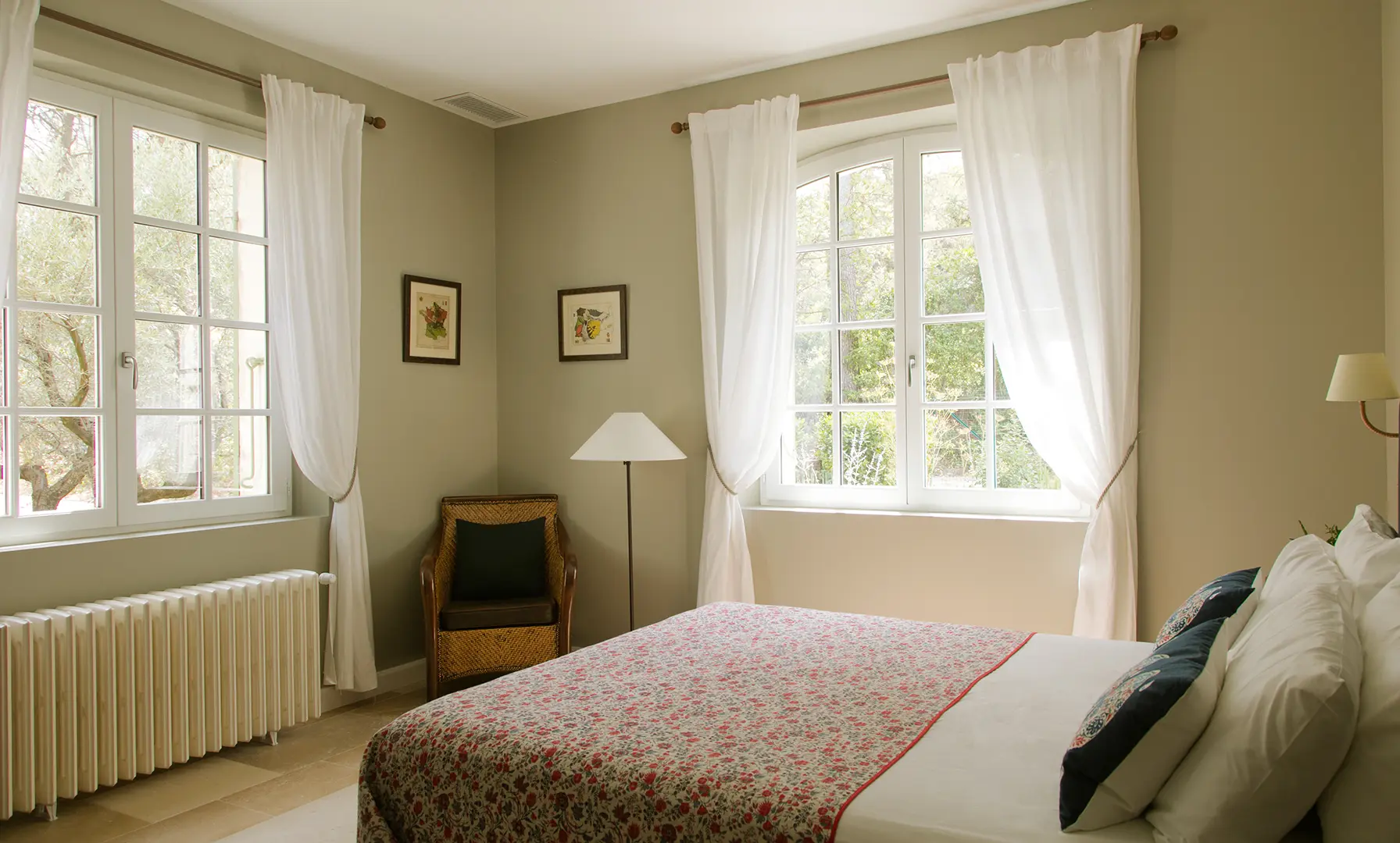 Une chambre confortable avec des murs couleur crème et une grande fenêtre avec des rideaux blancs. Le lit est orné d'une courtepointe fleurie et d'oreillers décoratifs. Une chaise en osier et un lampadaire sont situés dans le coin, avec des œuvres d'art encadrées sur les murs.