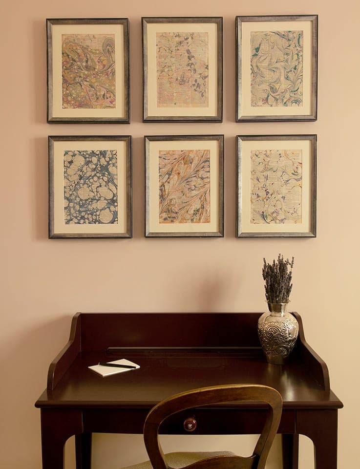 Un petit bureau en bois avec une chaise est placé contre un mur beige. Au-dessus du bureau se trouvent six œuvres d'art encadrées aux motifs variés. Un vase décoratif contenant de la lavande séchée est posé sur le bureau, à côté de quelques cartes vierges.