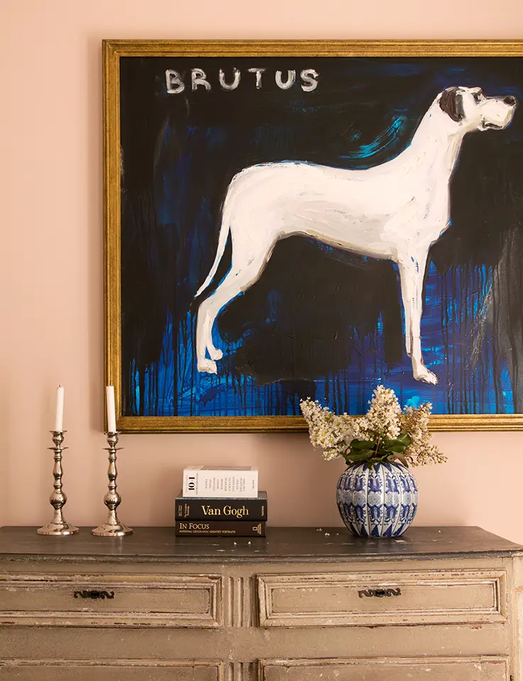 Sur un mur est accroché un tableau représentant un chien blanc sur lequel est écrit "BRUTUS". Au-dessous se trouve une commode en bois avec des bougeoirs en argent, des livres et un vase bleu et blanc avec des fleurs.