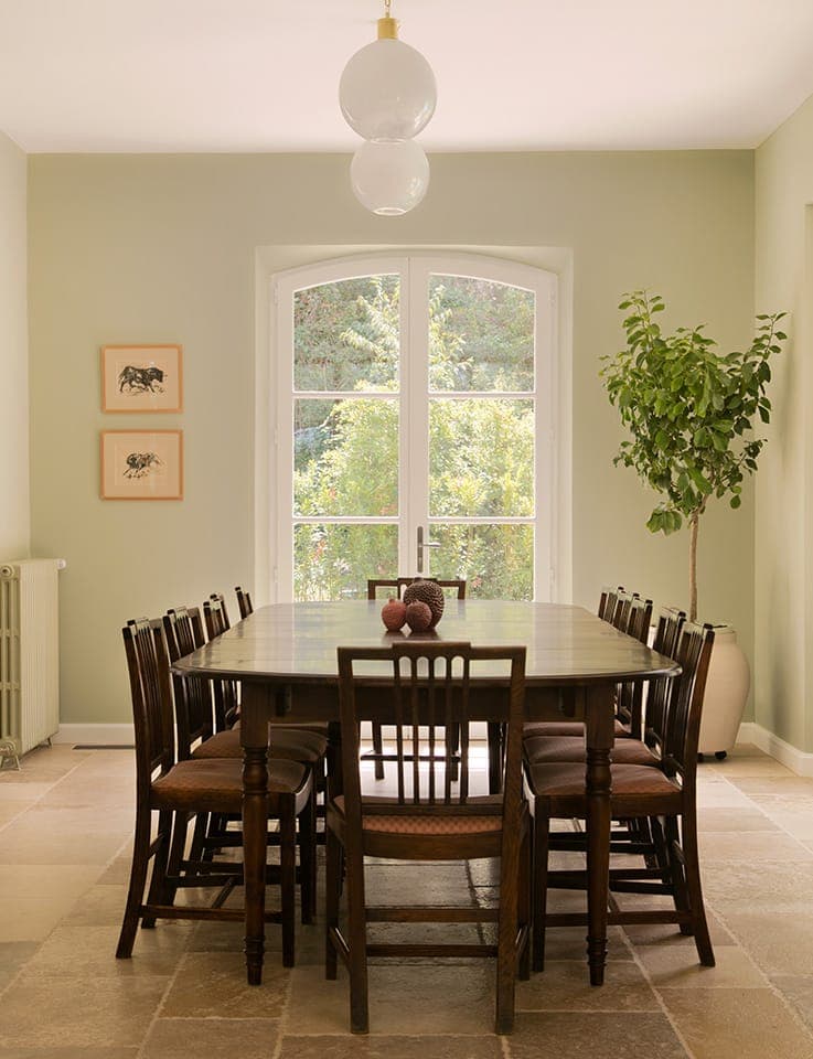 Une salle à manger avec une table en bois et huit chaises, éclairée par une suspension. Deux petites œuvres d'art encadrées sont accrochées sur un mur de couleur claire. De grandes fenêtres cintrées laissent entrevoir la verdure à l'extérieur, et une plante en pot se trouve à proximité.