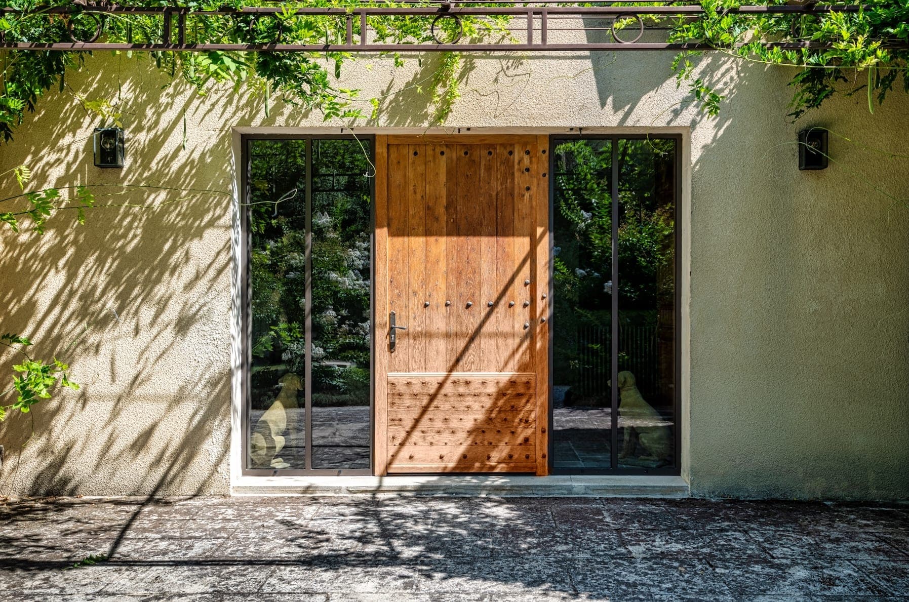 Une porte en bois avec des clous décoratifs est centrée entre deux grands panneaux de verre. Le mur est de couleur crème et des feuilles vertes projettent des ombres ludiques dessus. Le sol est carrelé en pierre et l'atmosphère générale est lumineuse et naturelle.