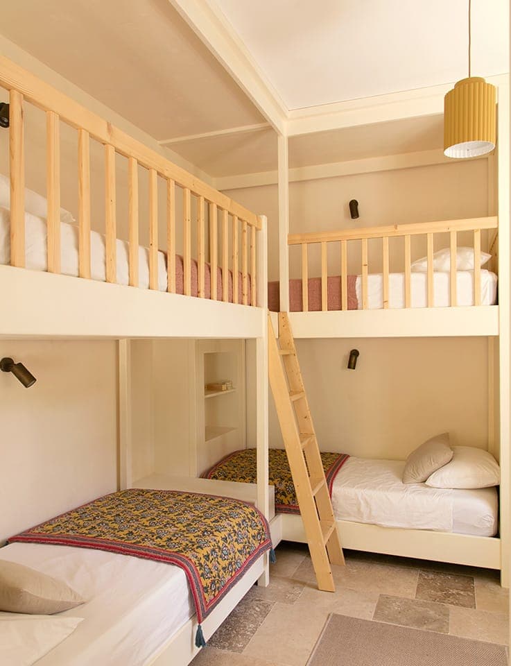 Une chambre confortable avec deux lits superposés en bois, chacun doté d'une couverture colorée et d'oreillers blancs. Une échelle en bois permet d'accéder aux lits supérieurs. La chambre a des murs de couleur crème, une lampe suspendue et de petites lampes murales.