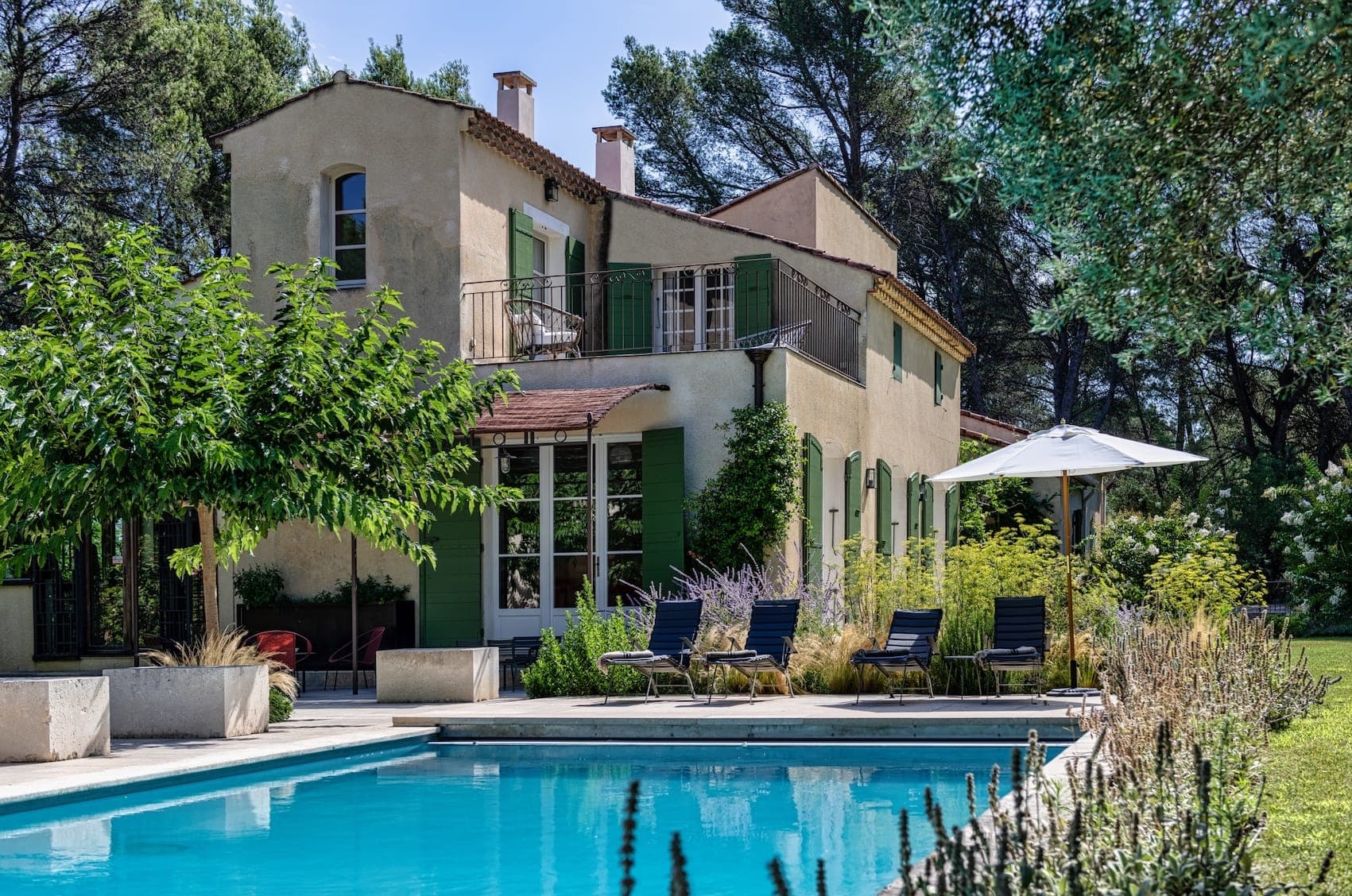 A Mediterranean-style villa with cream walls and green shutters, surrounded by lush greenery. It features a pool with several lounge chairs and a large umbrella in the backyard, under a clear blue sky.