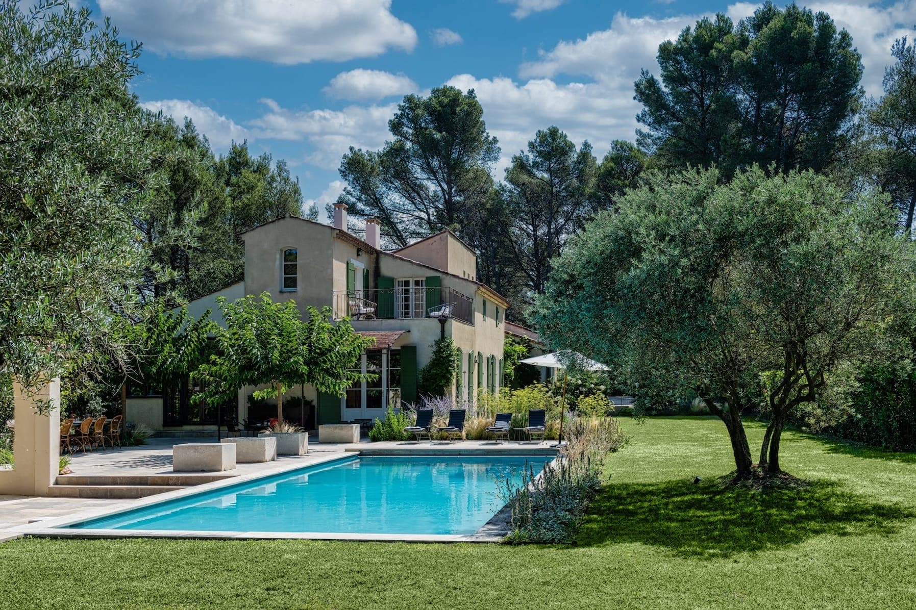 Une villa sereine entourée d'une végétation luxuriante et d'arbres dispose d'une grande piscine accueillante. La maison a un extérieur pâle avec de multiples fenêtres et patios. Le ciel est bleu vif avec des nuages épars, ajoutant à l'ambiance tranquille.